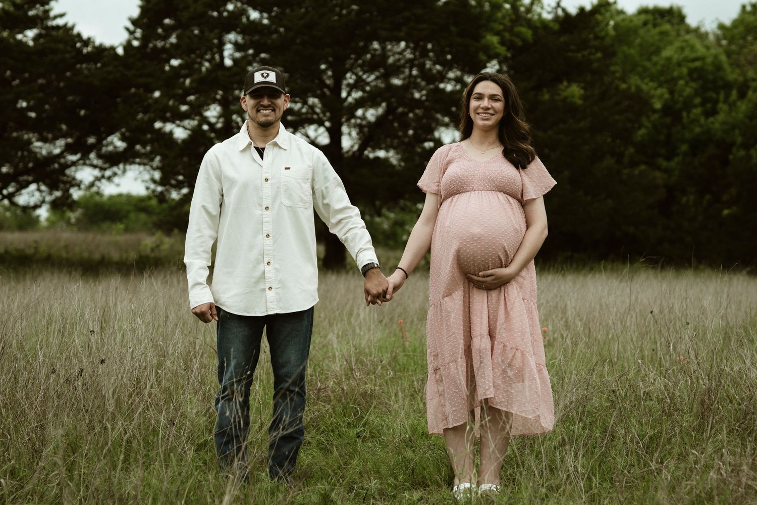 Expectant couple stands together in tall grass field wearing coordinating pink dress and white shirt.