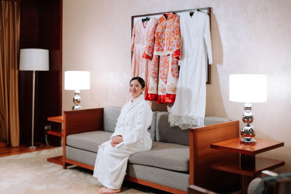 Wedding attire displayed on wall with person in white robe sitting on gray couch in hotel room.