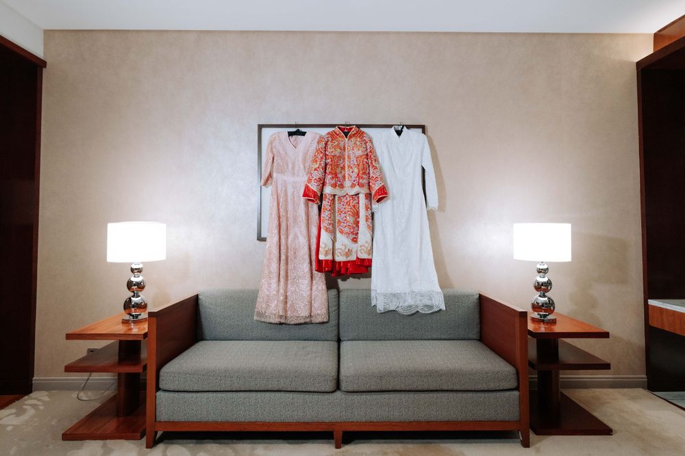 Traditional red and white wedding attire displayed on hangers above a gray couch with table lamps in a hotel room.