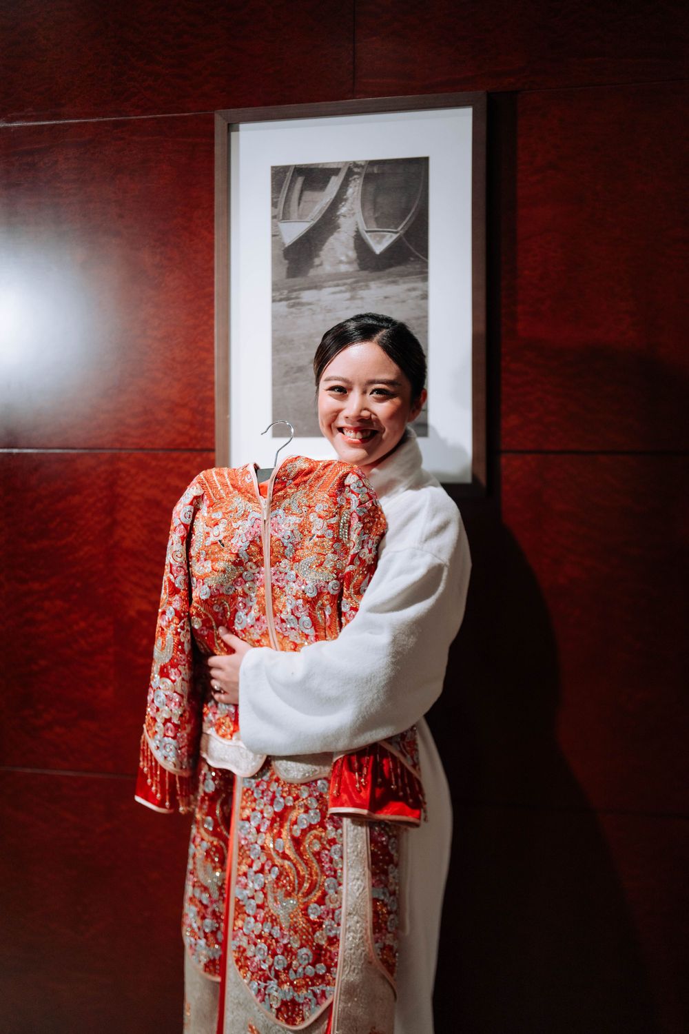 Person wearing white robe holding traditional red patterned wedding dress against dark wall.