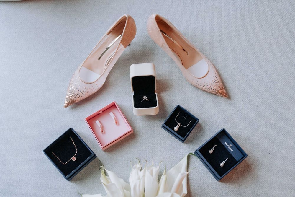Blush pink wedding shoes arranged with jewelry boxes and feathers in flat lay composition.