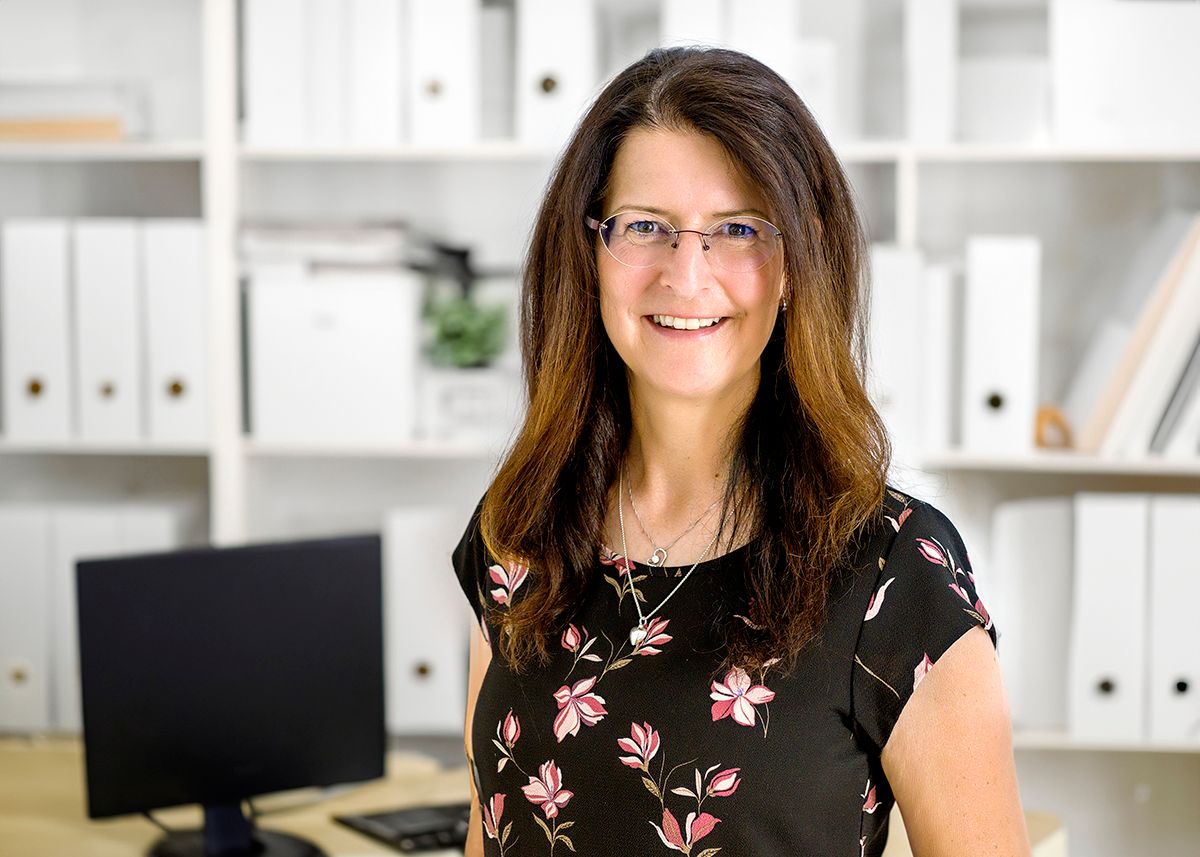 Businessfoto einer Frau mit langen Haaren und Brille vor einer hellen Bürowand