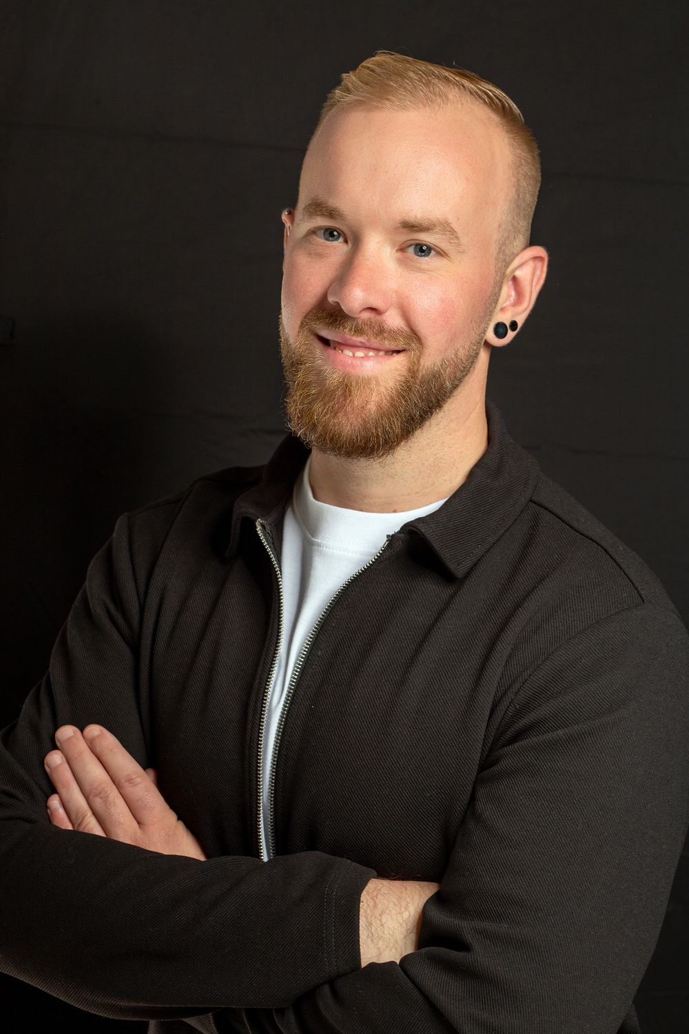 Professional studio portrait of a smiling person with a blonde beard wearing a black jacket and white shirt against dark background.