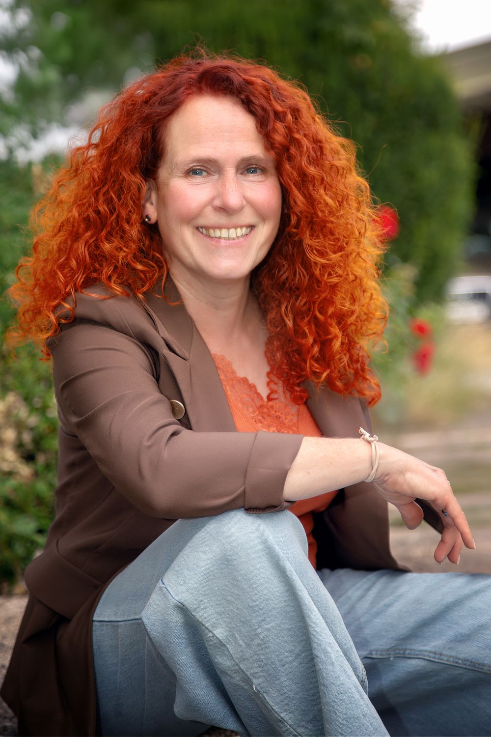 Smiling woman with curly red hair wearing a brown blazer , sitting outdoors in a garden setting. Bewerbungsfoto Gartencentermitarbeiterin