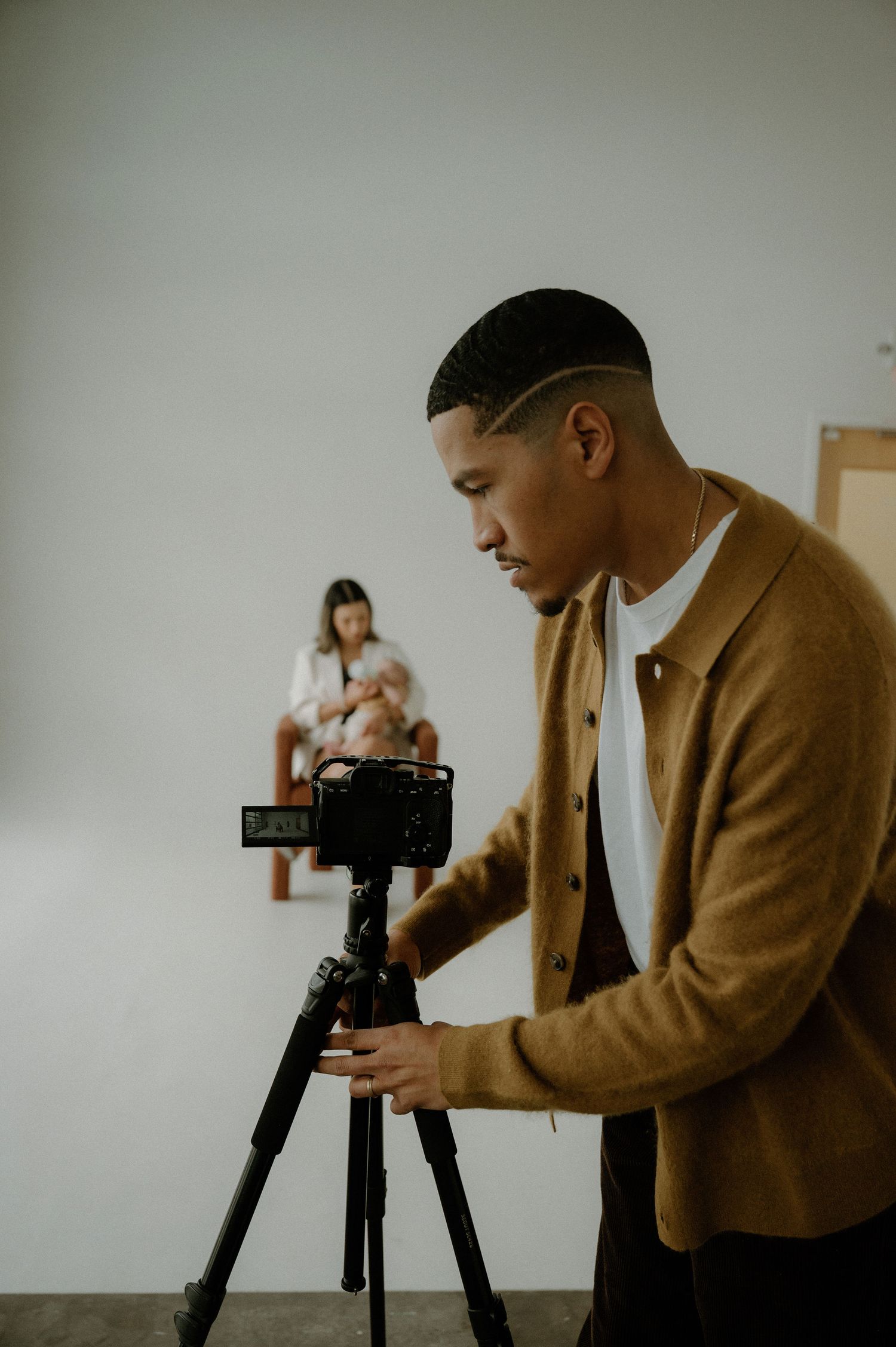 A photographer in a brown cardigan adjusts a camera on a tripod while a model sits in soft lighting at a photo shoot.