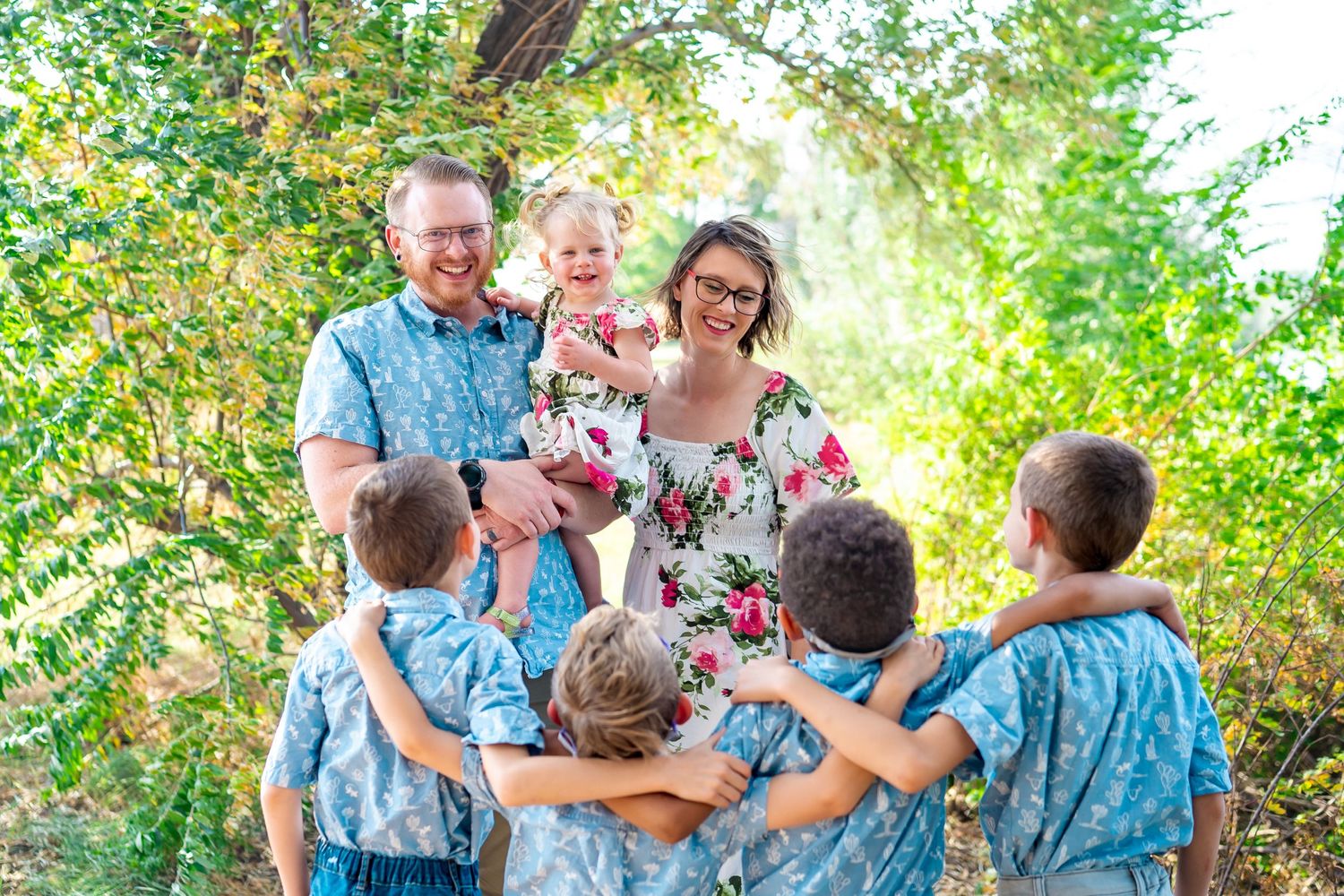 Family group wearing coordinating blue outfits embraces in a sunny outdoor setting.
