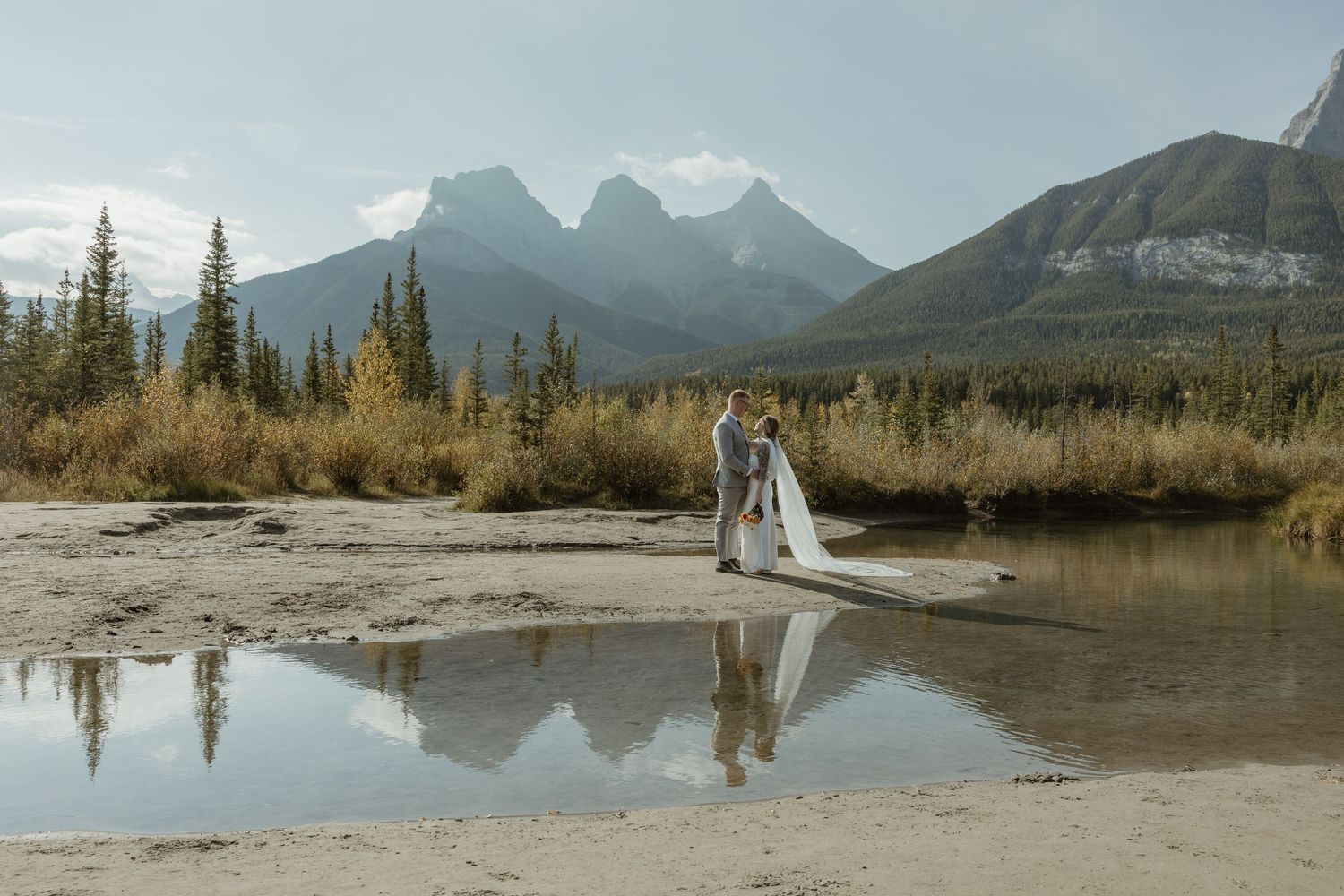 Bride and groom embrace near a reflective mountain stream in Bismarck, ND with the mountain peaks in the background.