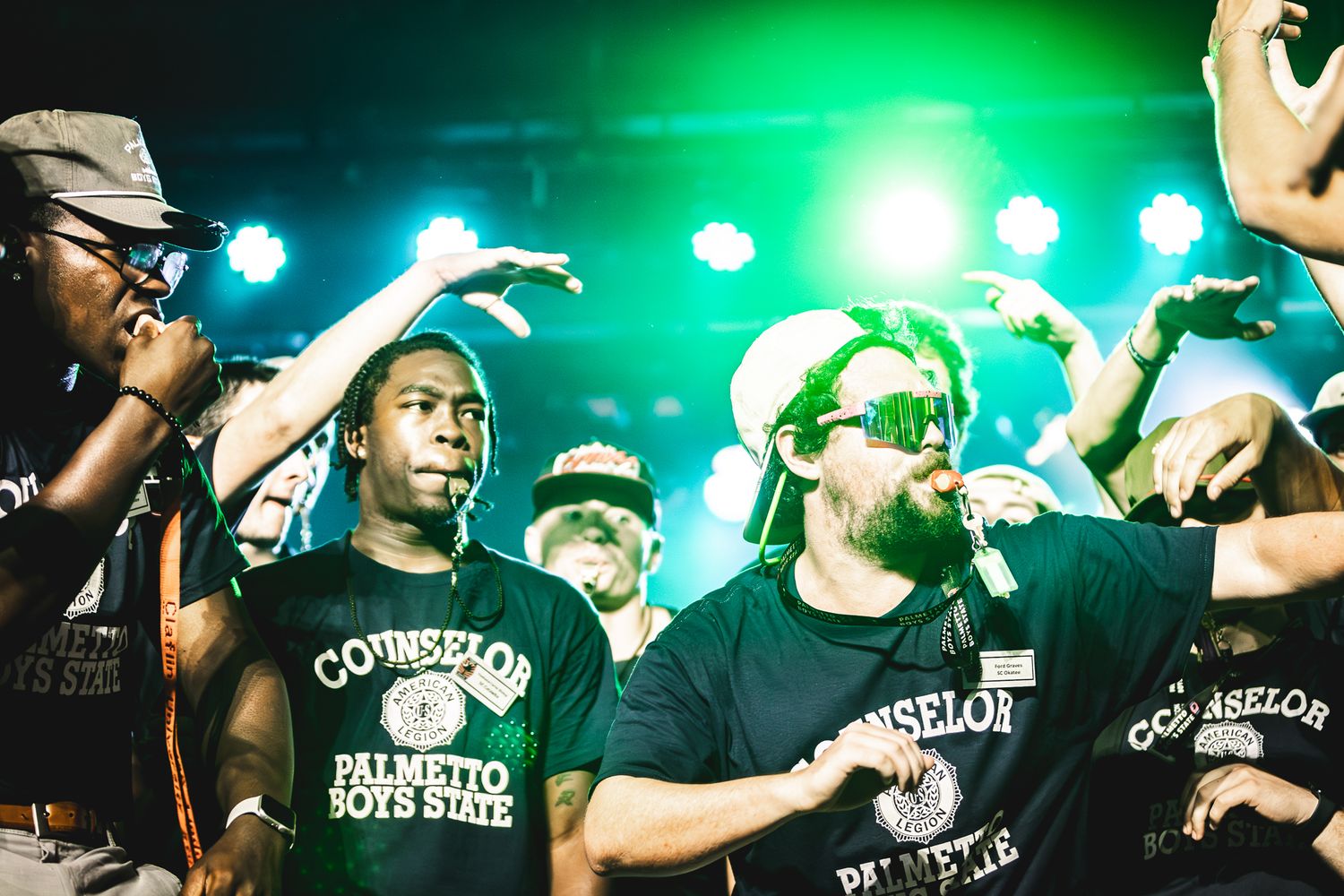 Camp counselors in dark Palmetto Boys State shirts raise their hands and cheer together under green stage lights.