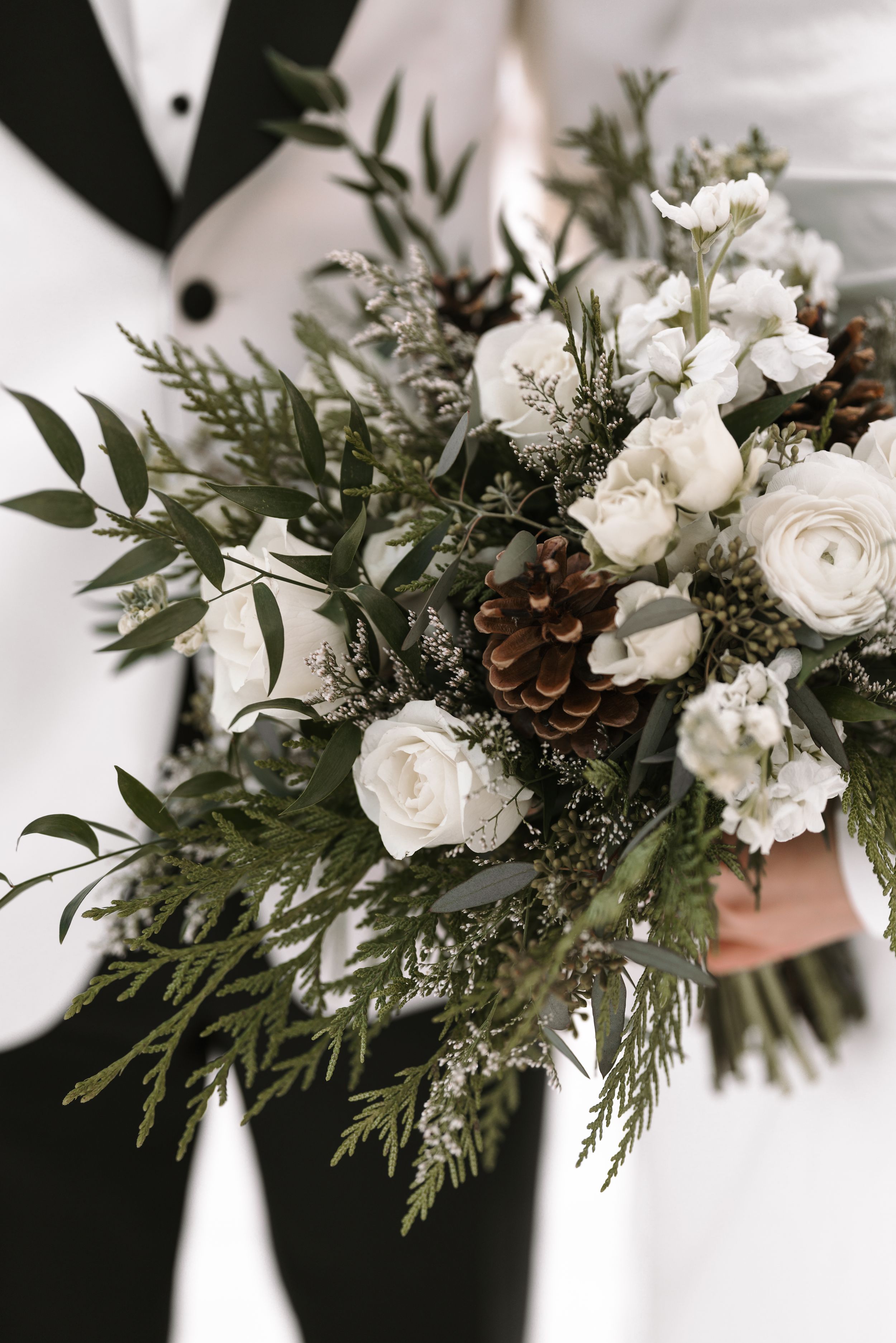 White ranunculus and pine cone winter wedding bouquet with green eucalyptus foliage and evergreen sprigs.