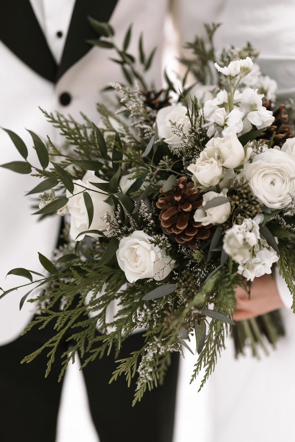 White ranunculus and pine cone winter wedding bouquet with green eucalyptus foliage and evergreen sprigs.