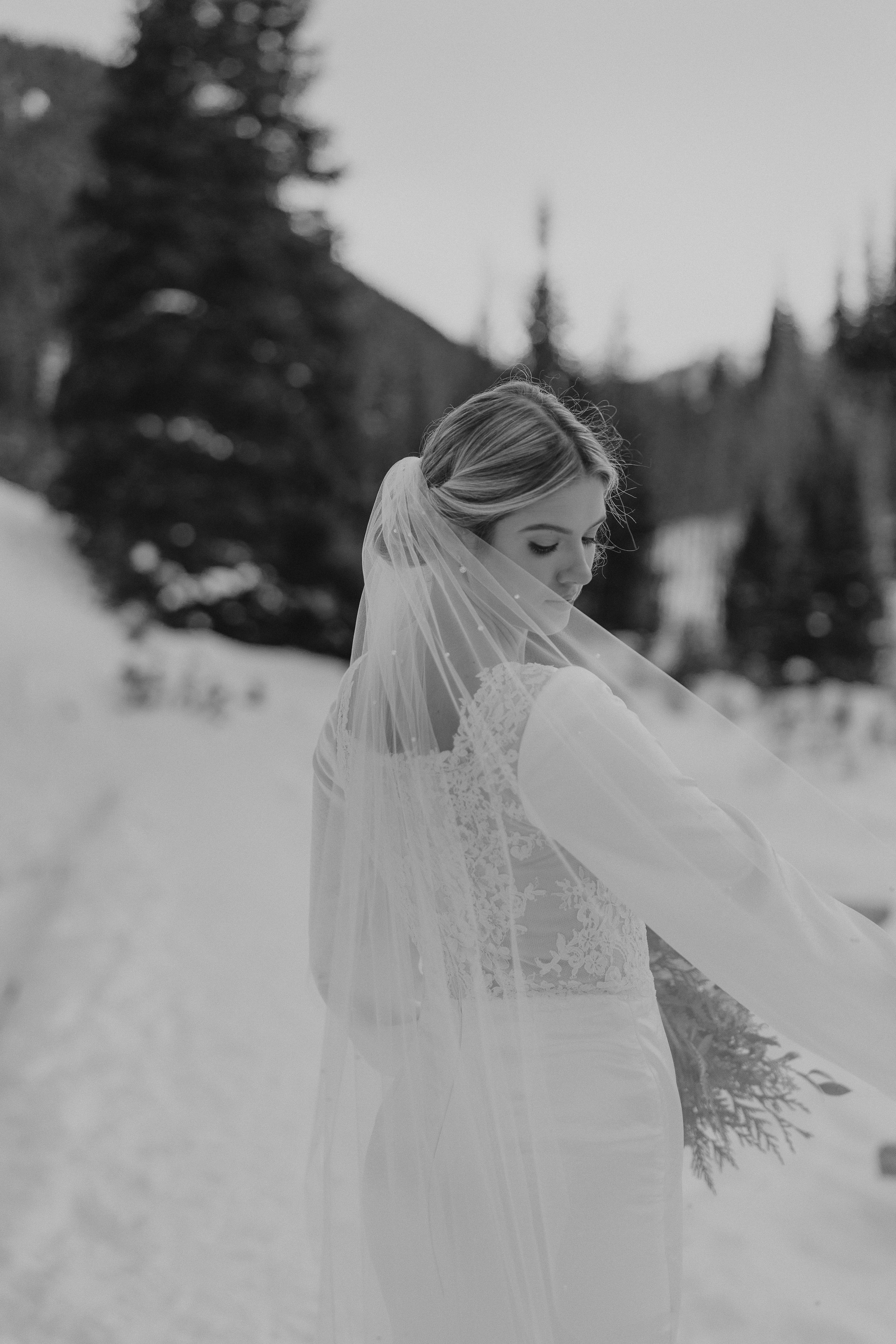A flowing white wedding veil catches the breeze against a snowy mountain backdrop during a winter photoshoot.