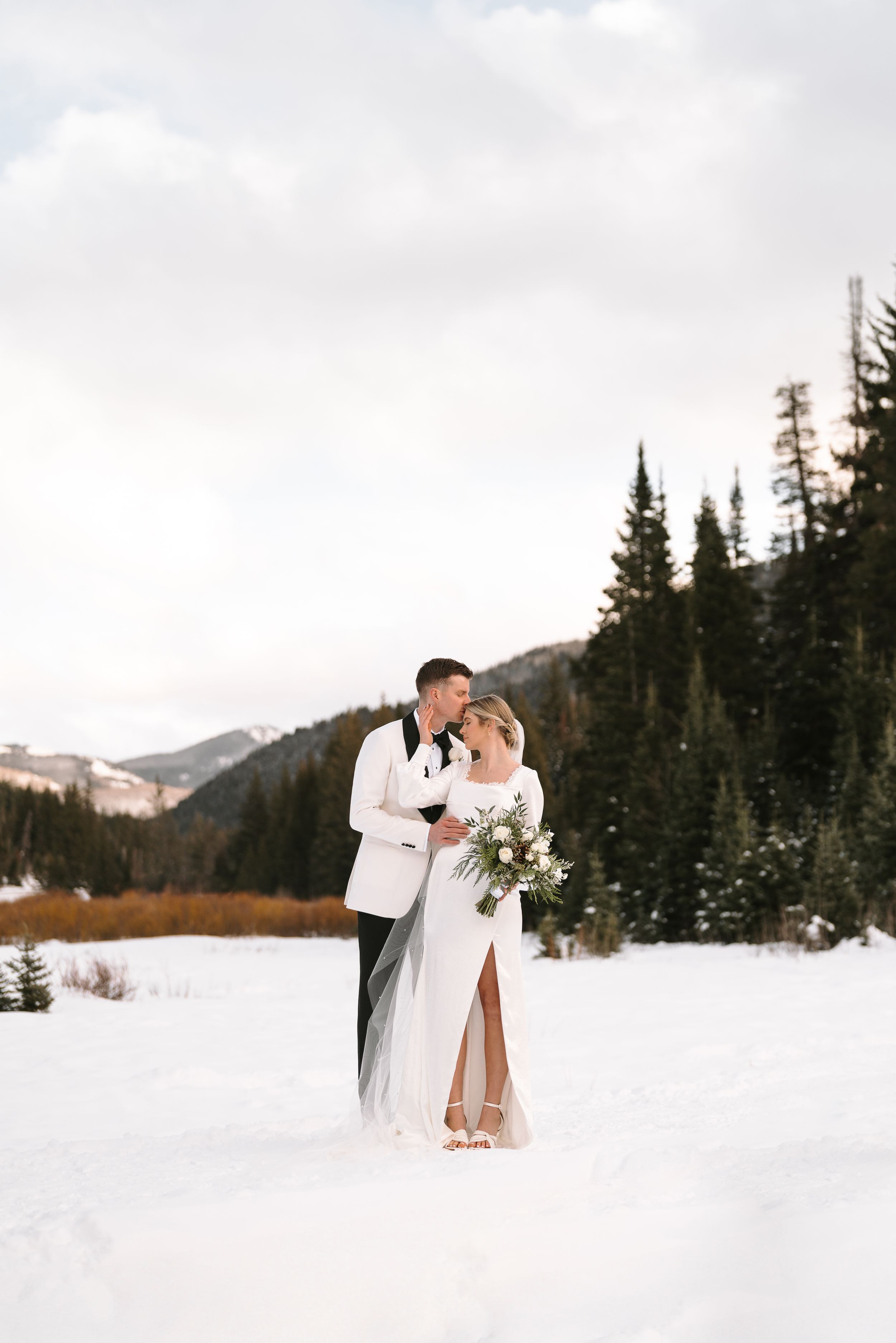 A couple shares a romantic moment in a snowy winter landscape surrounded by evergreen trees and mountains.