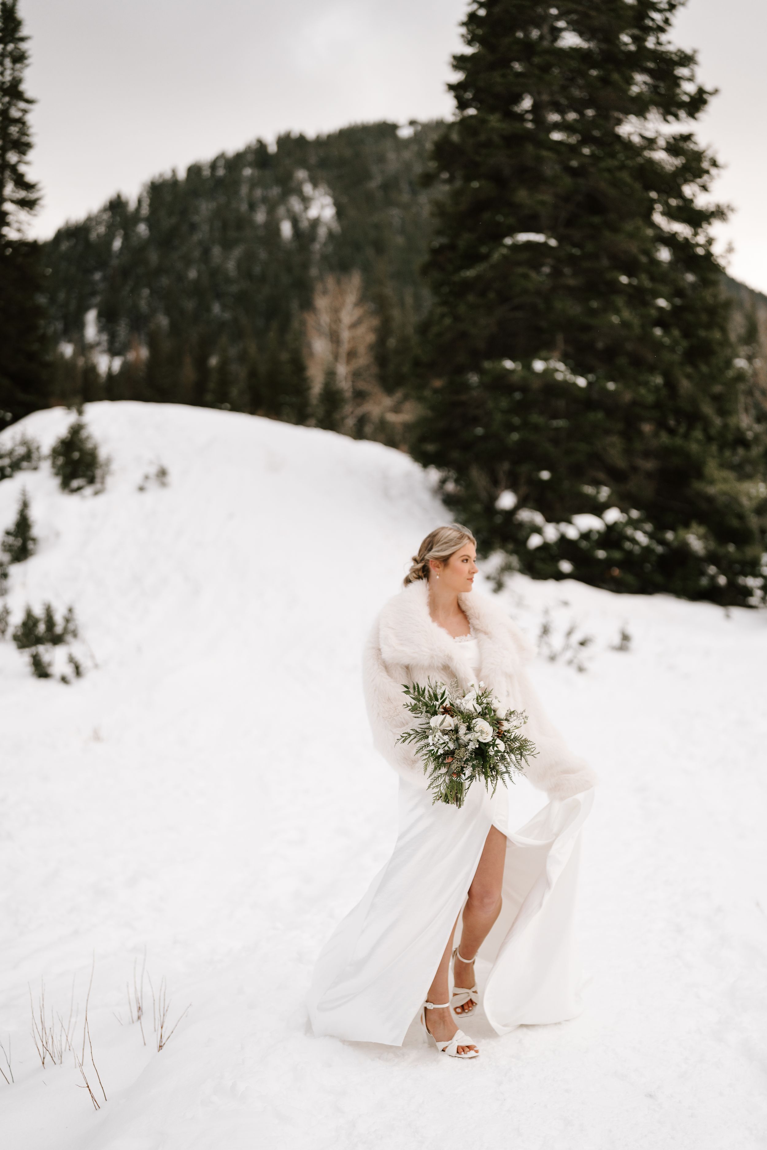 A winter bride in white dress gracefully poses in snowy mountain landscape with evergreen trees.