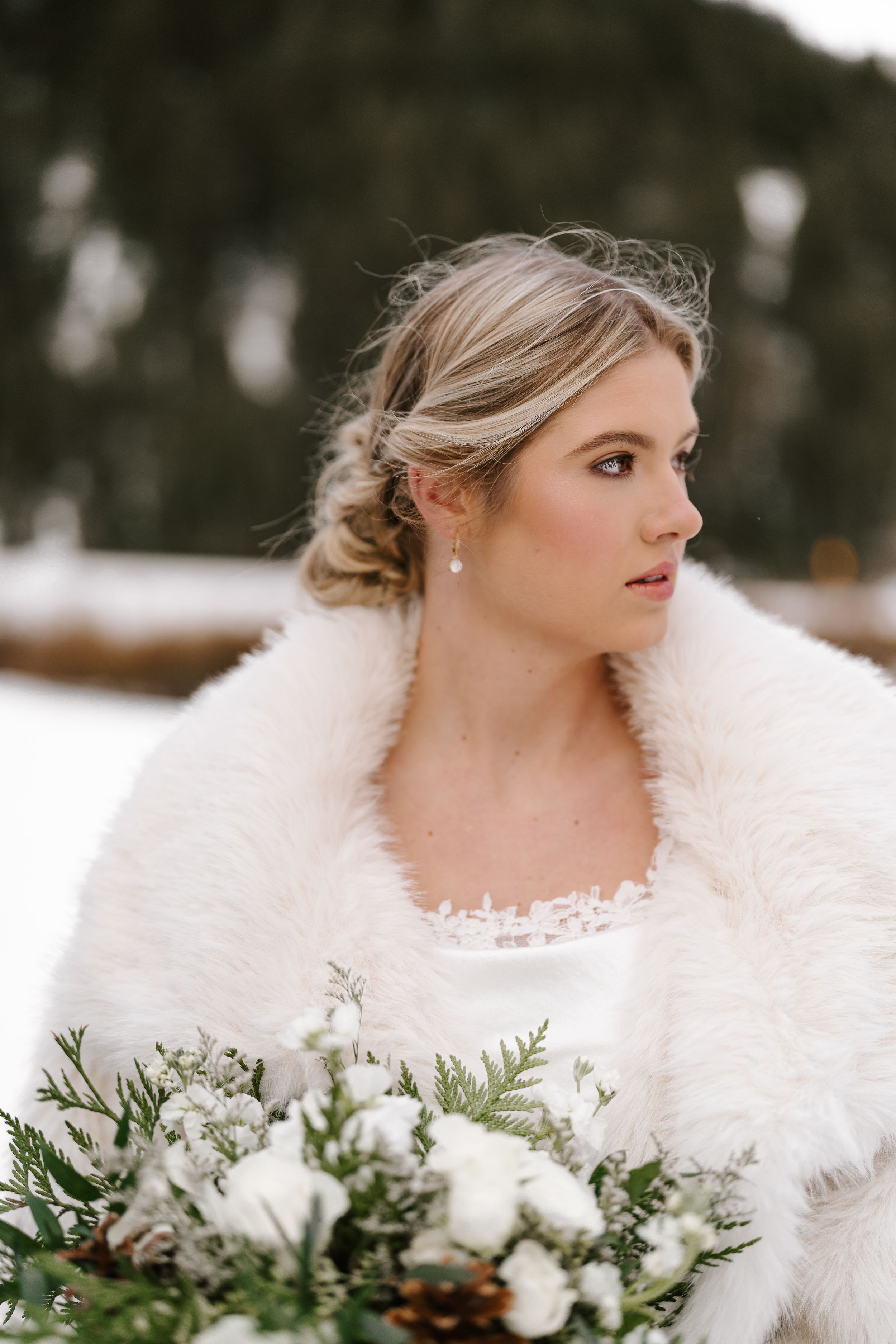 Winter bride in white fur stole holds evergreen bouquet with white flowers against snowy backdrop.