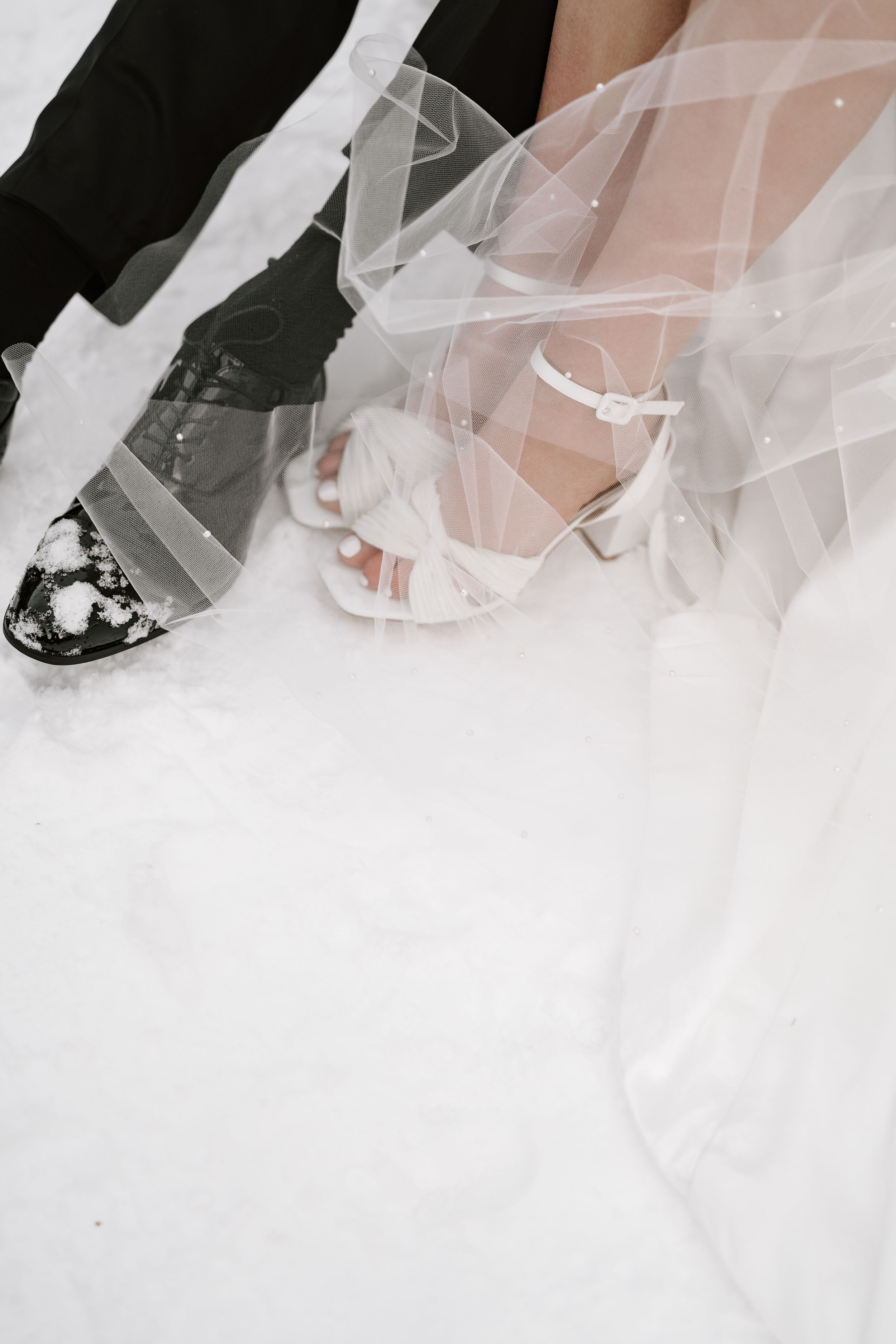 Close-up of dress shoes and the hem of a white wedding dress trailing on the floor in black and white.