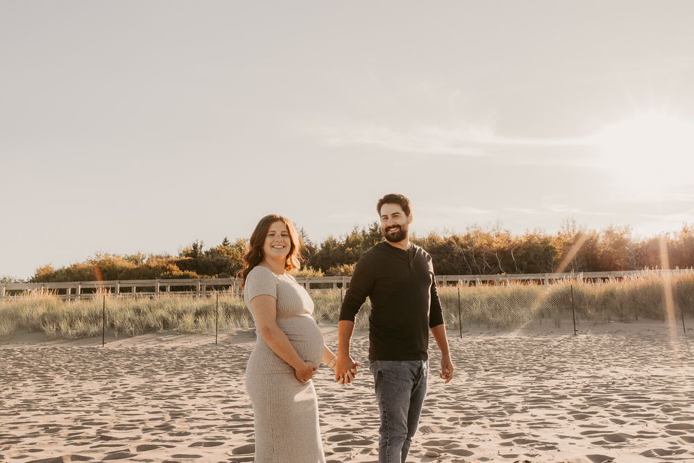 Pregnant couple holding hands on sandy beach at golden hour, woman in gray maternity dress smiling over shoulder.