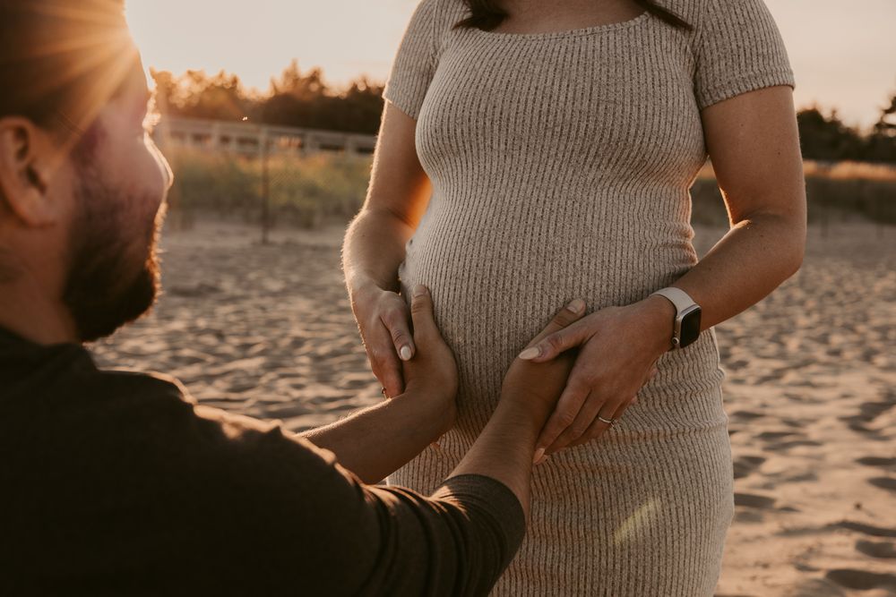 Pregnant woman in beige dress touched by partner on sunny beach at golden hour.