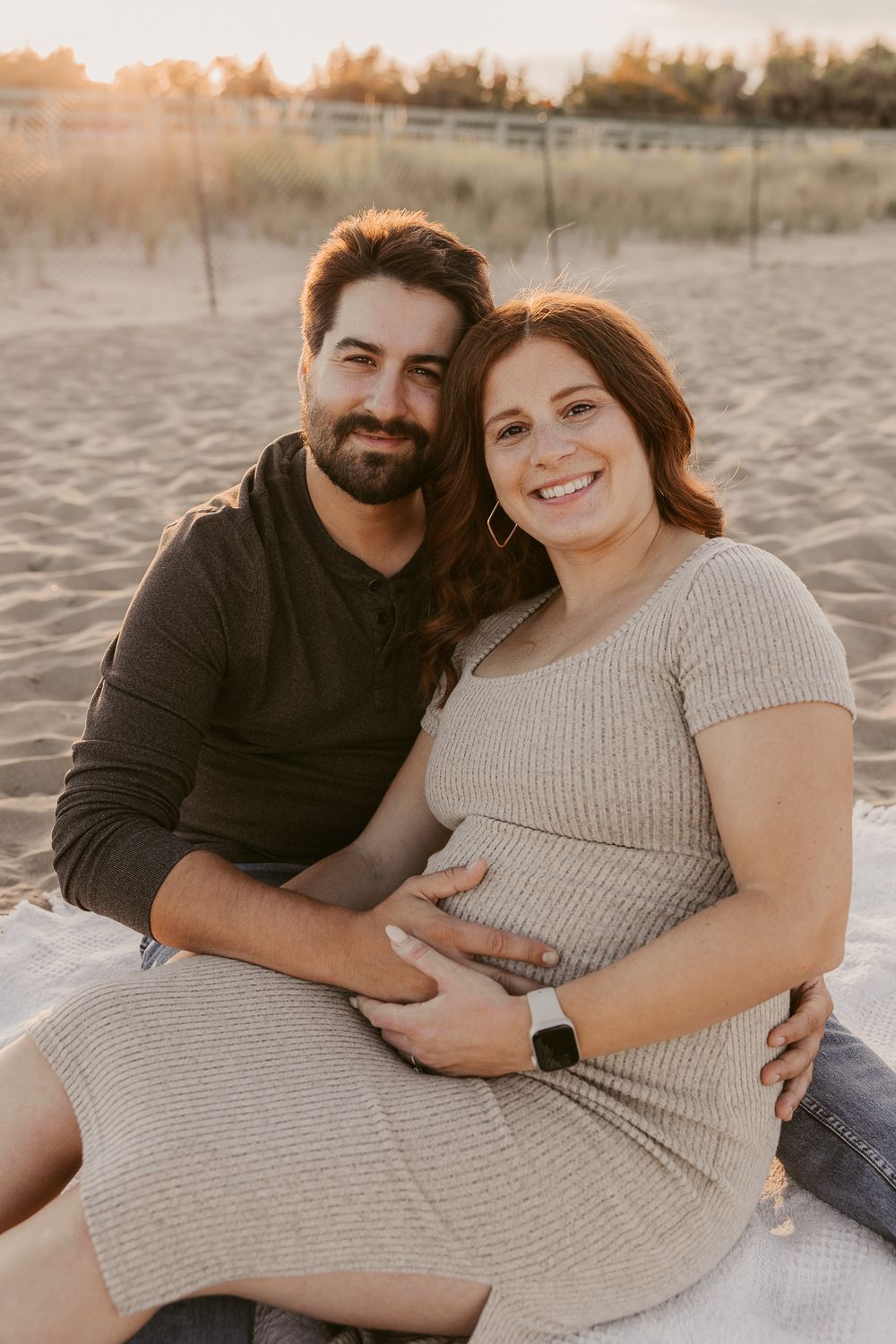 Smiling pregnant couple sitting on sandy beach at golden hour, man embracing woman who cradles baby bump.