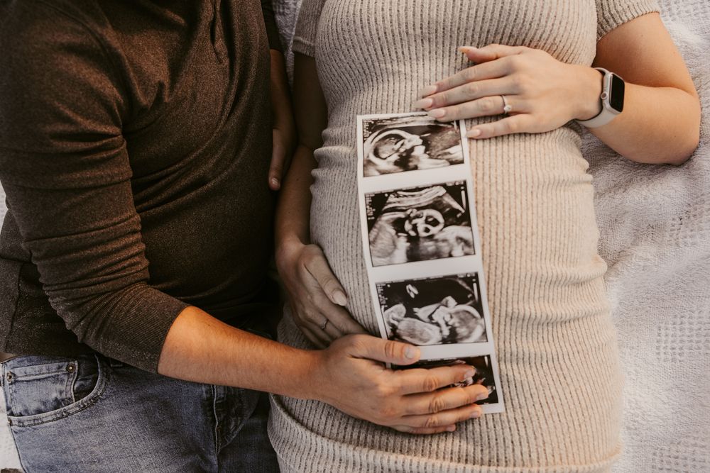 Pregnant couple holding ultrasound photos while man gently touches baby bump in intimate maternity photo.