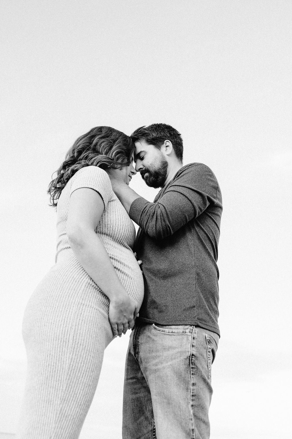 Black and white maternity photo of a couple tenderly touching foreheads outdoors, celebrating their pregnancy together.