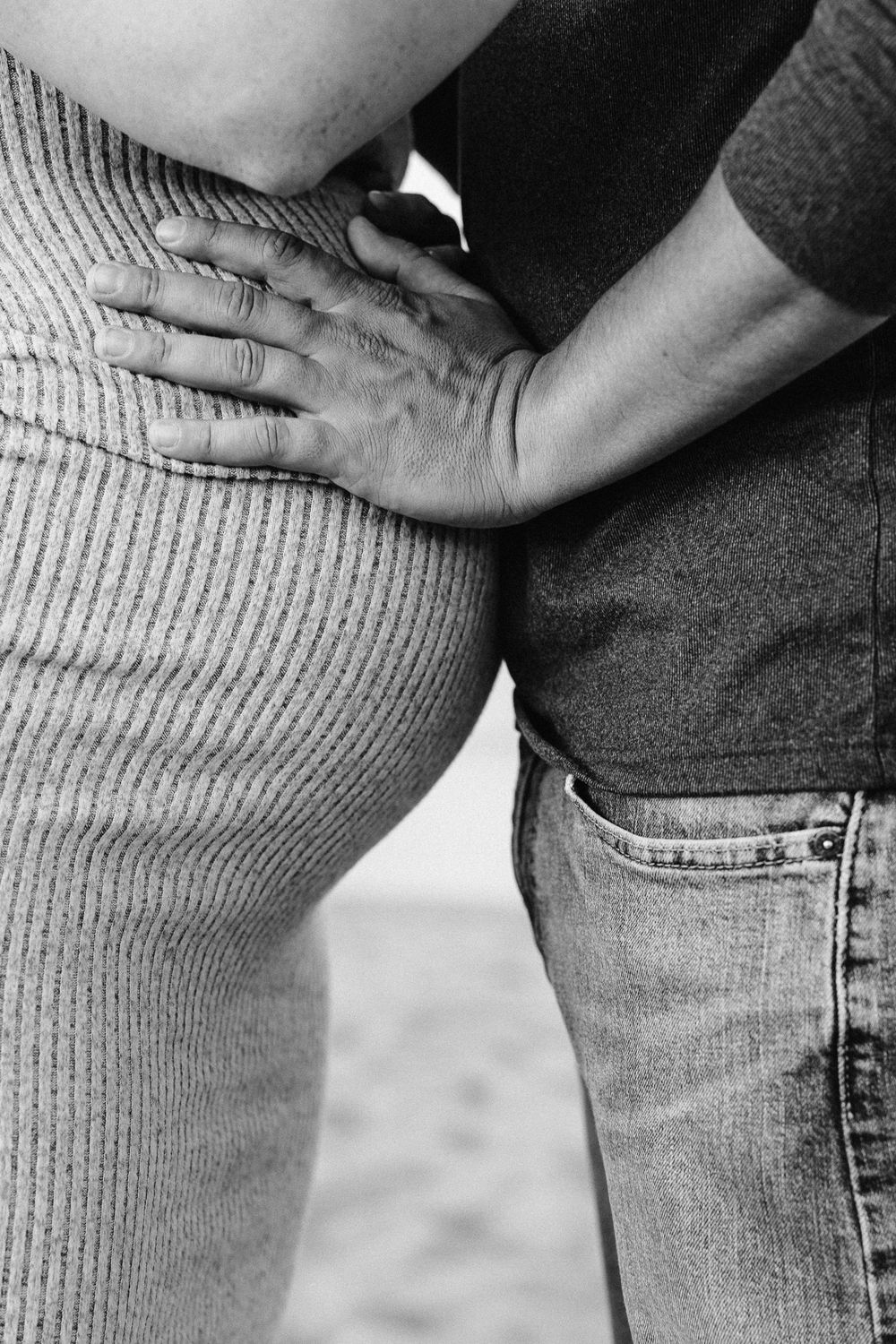 Black and white photo of a person placing hand on pregnant woman's baby bump, wearing knit dress and jeans.