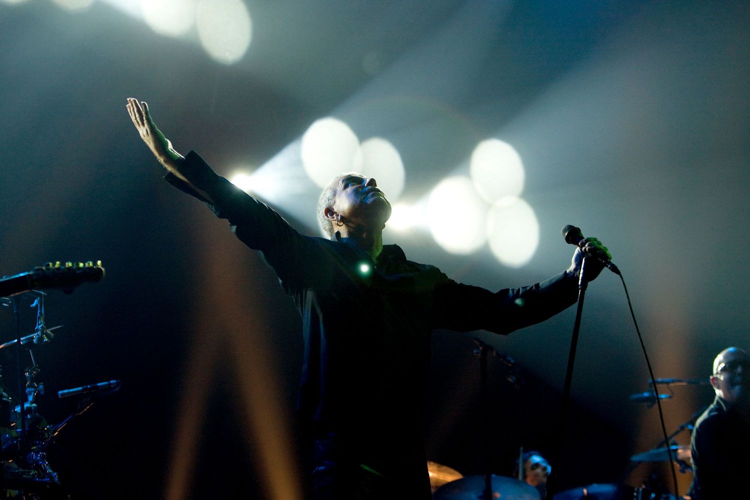 Concert performer silhouetted against bright bokeh lights raises arms dramatically on stage.