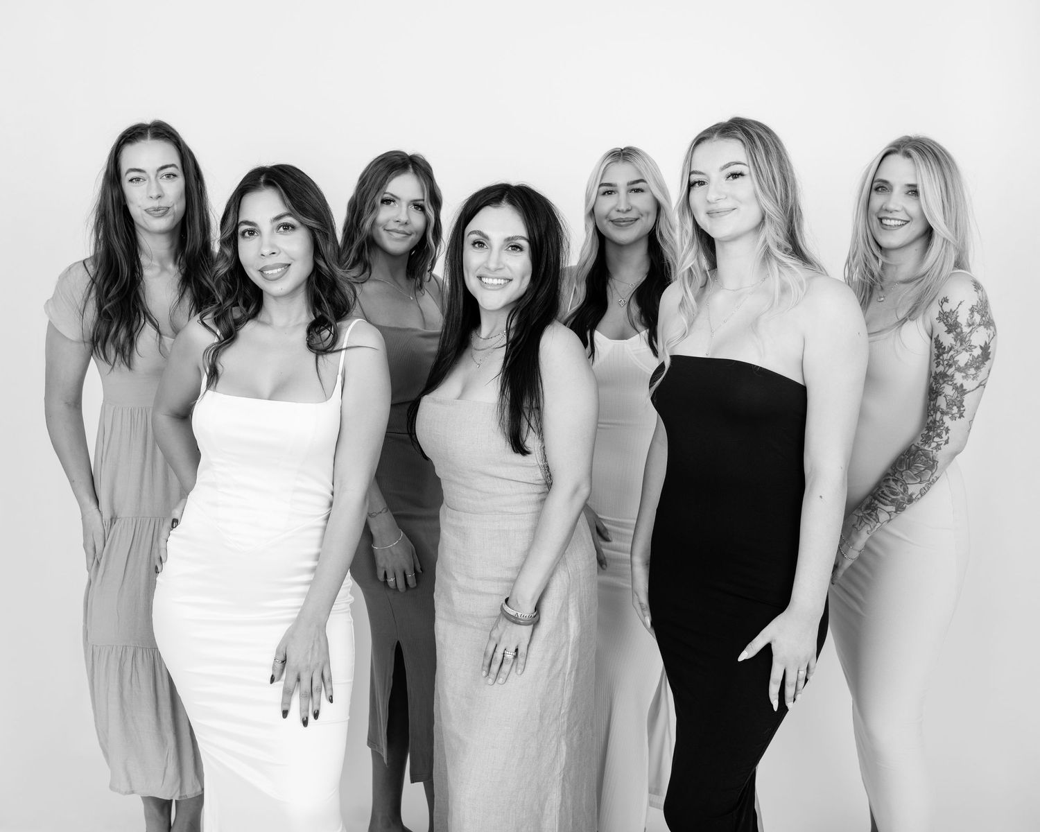 Black and white portrait of a group of seven women standing together in elegant dresses posing for a formal photo.
