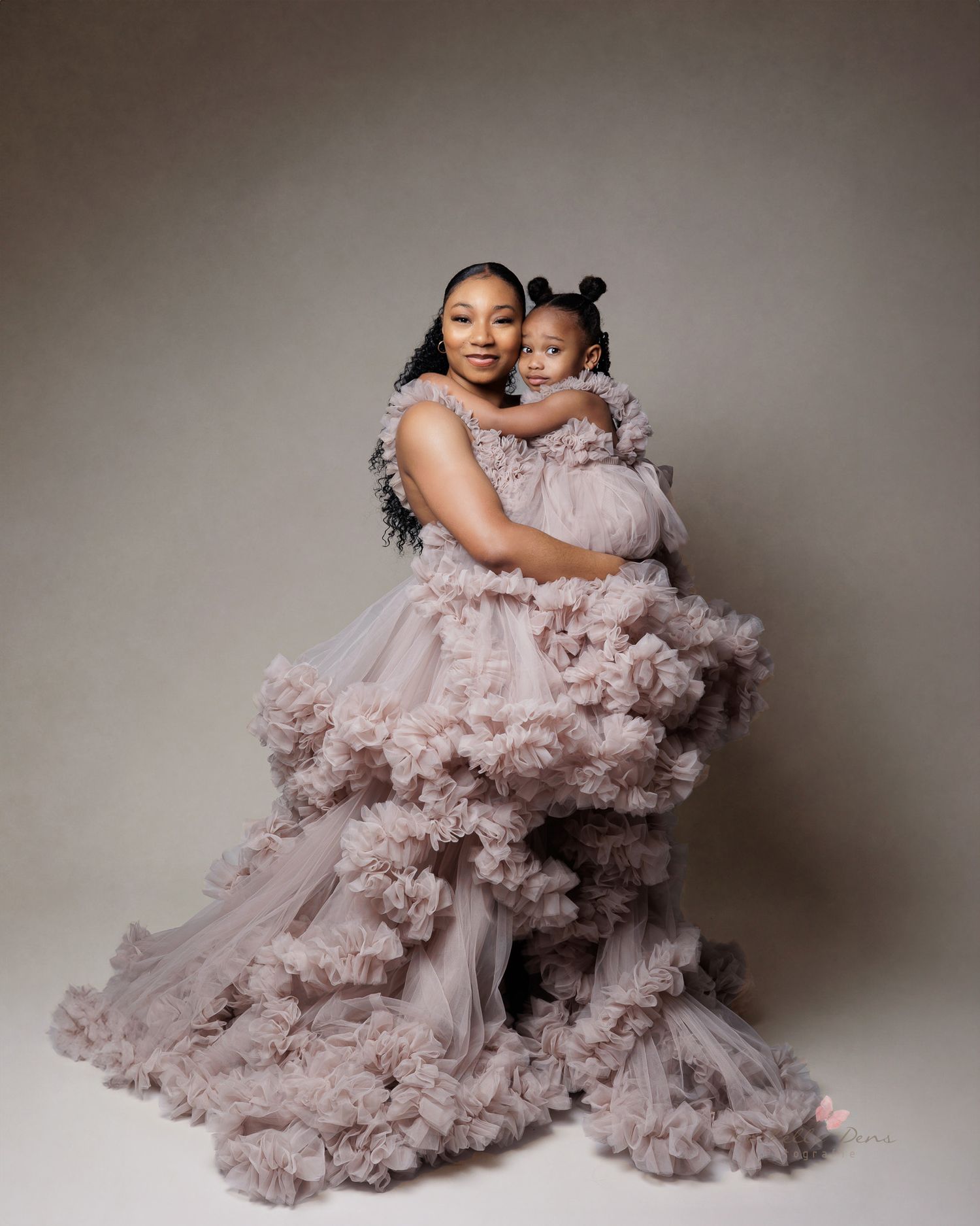 Mother and child embracing in flowing pink tulle dresses against a neutral studio backdrop.