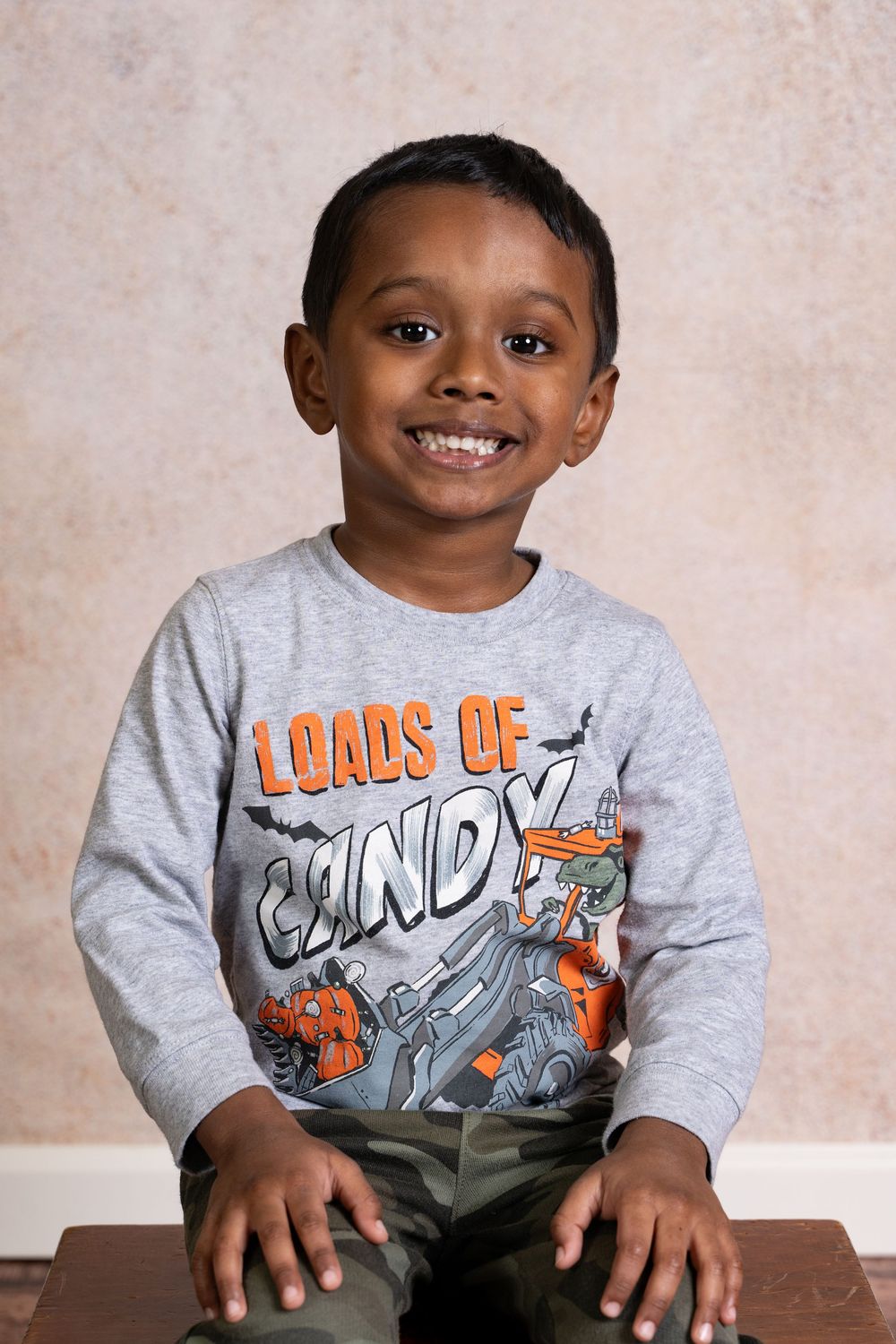 A young child wearing a gray sweatshirt with orange text sits at a wooden table smiling at the camera.