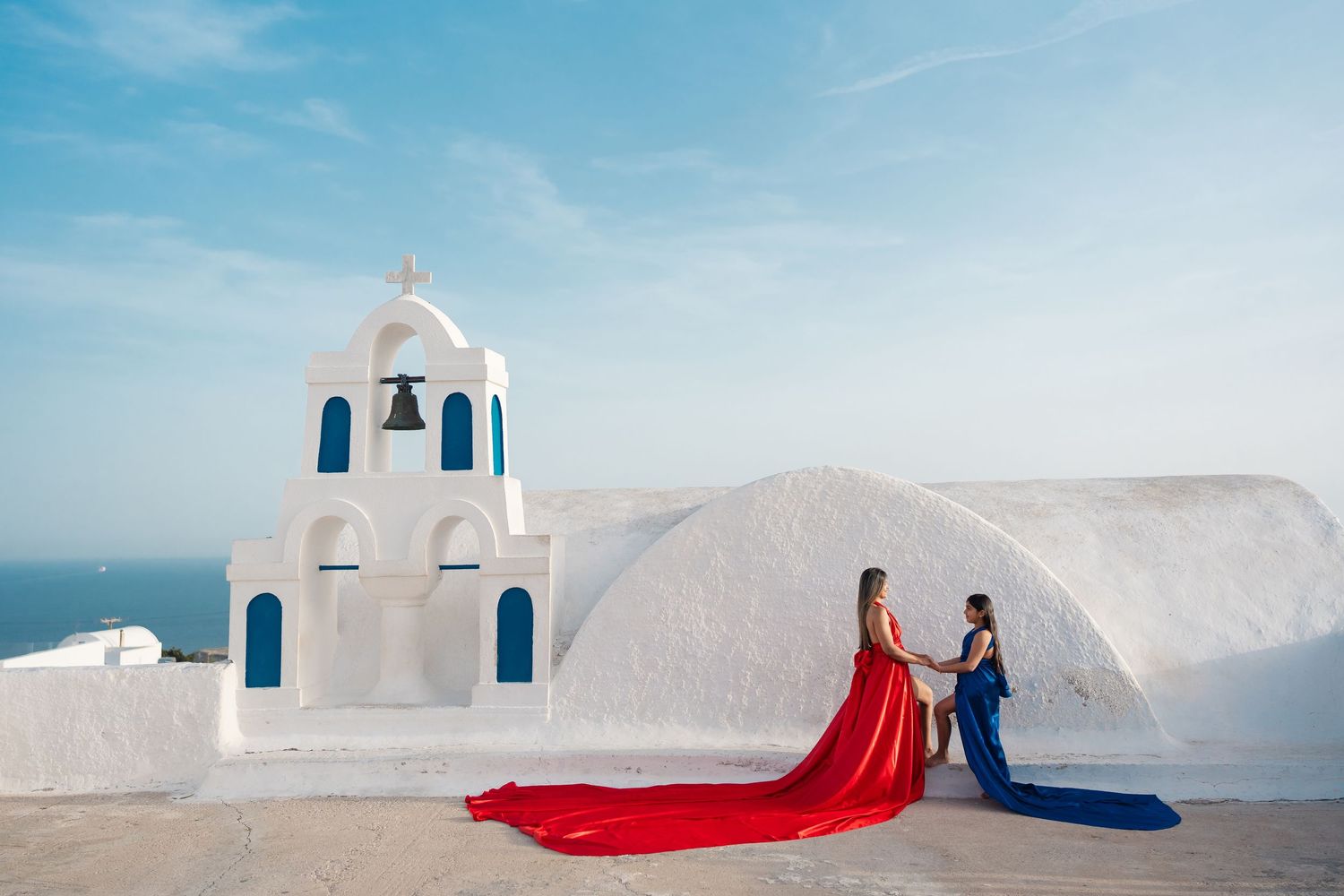 Red and blue flowing dresses create dramatic scene in front of white Greek church with blue domes.