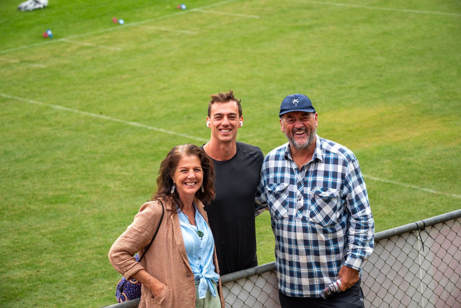 Three people smile together on green hillside near sports field.