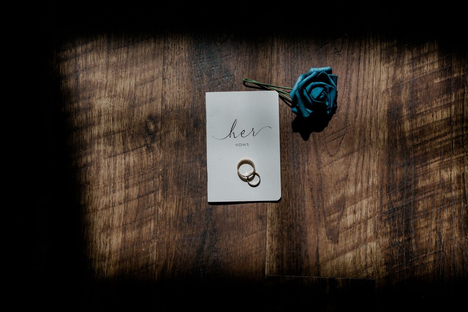 Wedding rings and a 'Love' card displayed on rustic wooden surface with blue rose accent in dark moody lighting.