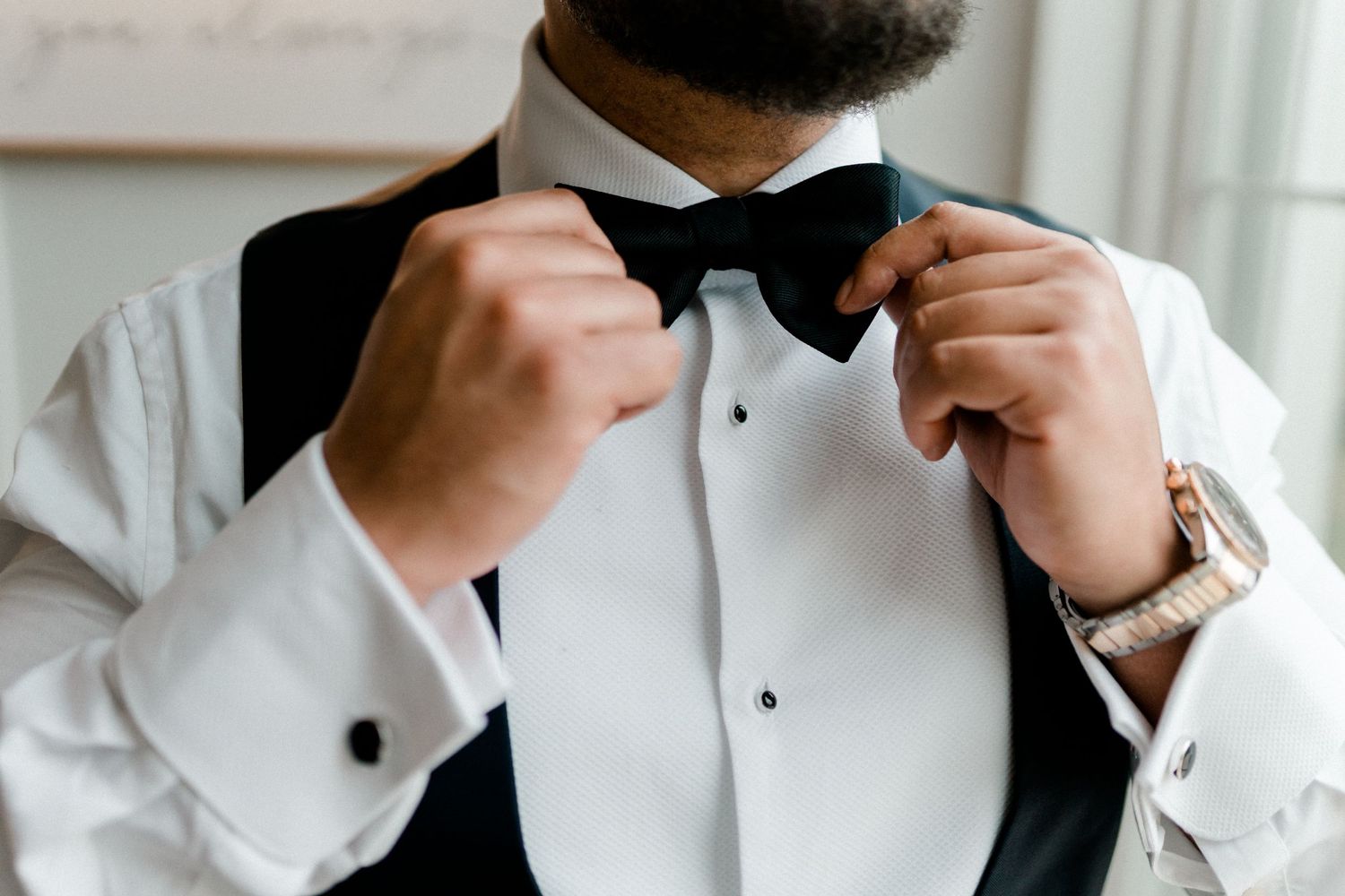Person adjusts black bow tie while wearing formal white tuxedo shirt and vest.