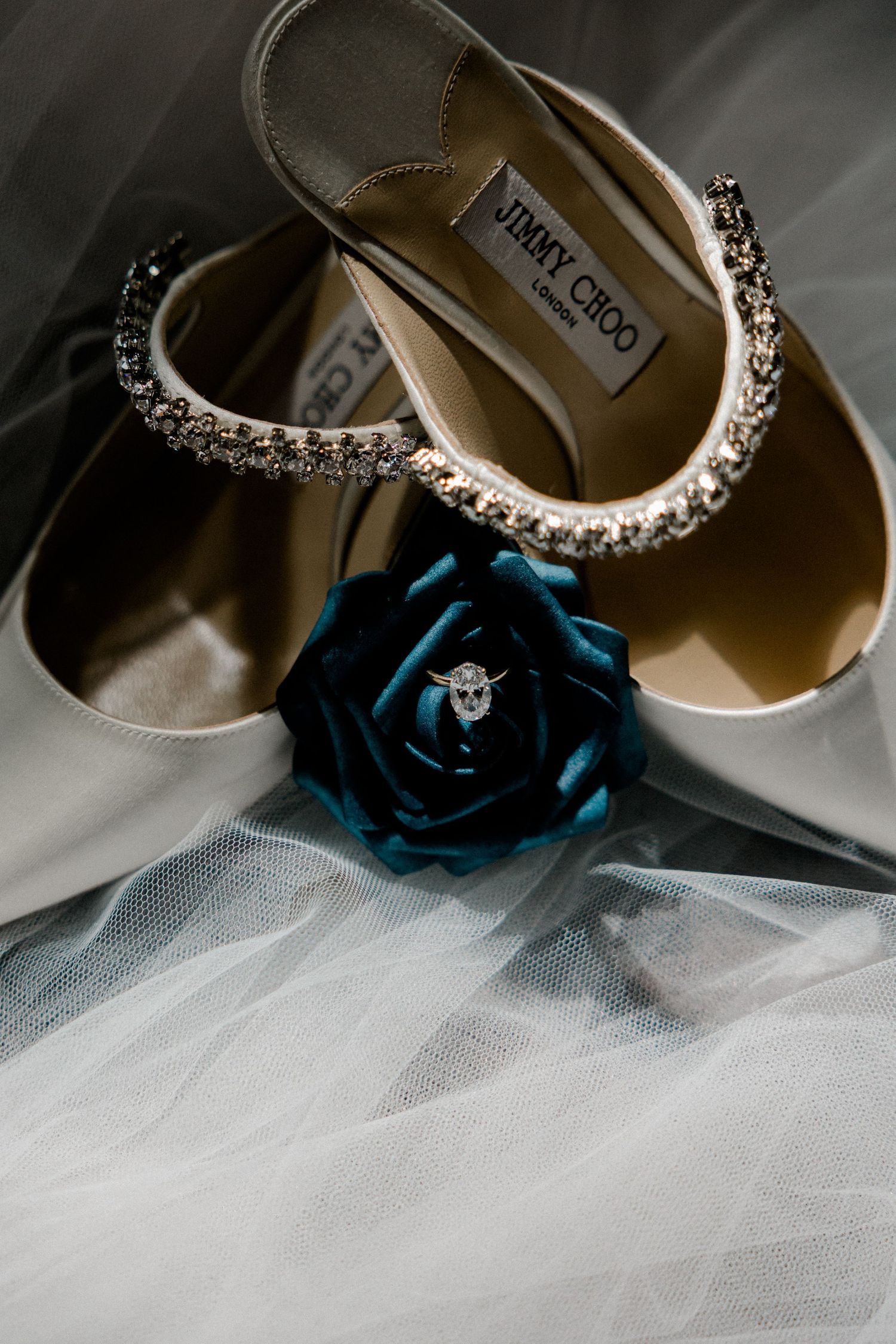 Elegant Jimmy Choo bridal shoes with crystal embellishments displayed with a teal blue rose on white tulle fabric.
