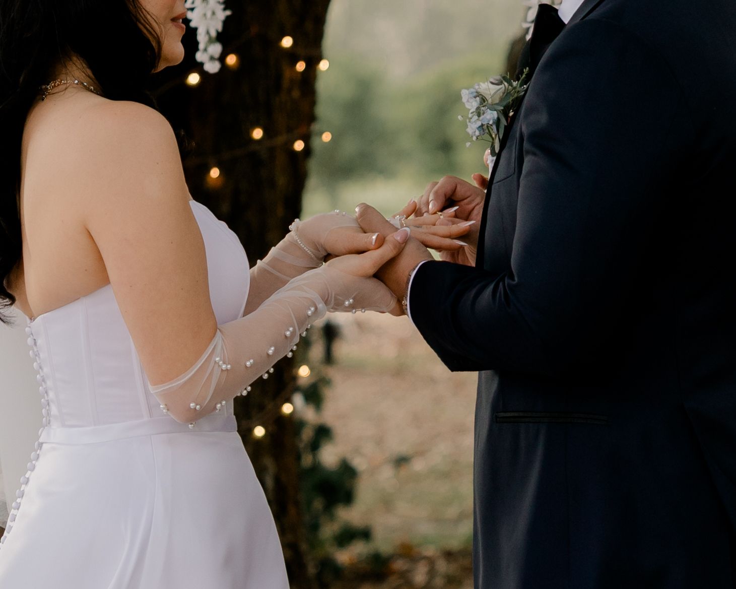 Close-up of hands exchanging wedding rings during outdoor ceremony with twinkle lights in background.