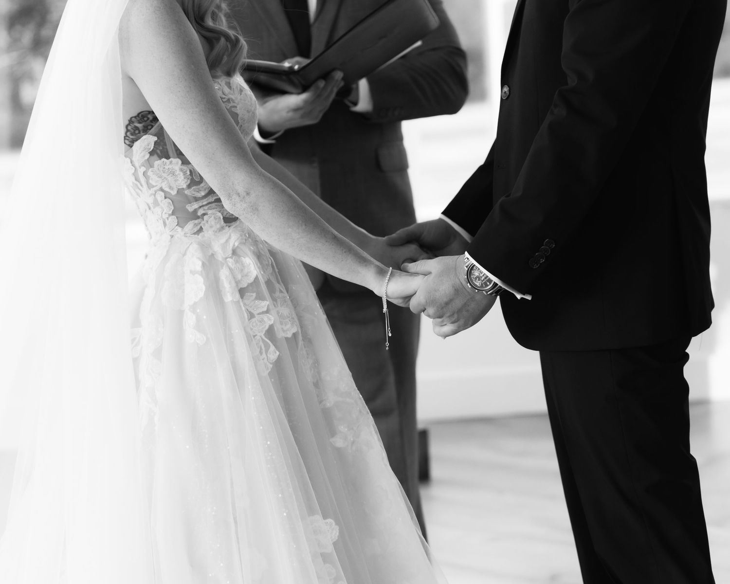 A bride and groom hold hands during their wedding ceremony while surrounded by guests in formal attire.