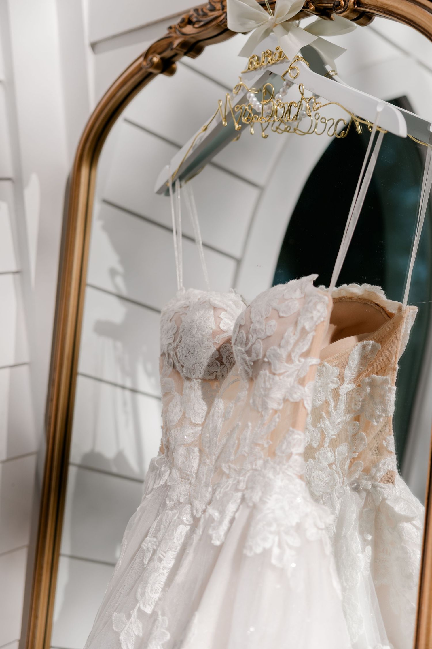White lace wedding dress hangs elegantly on a hanger against a white wall with ornate gold-framed mirror.
