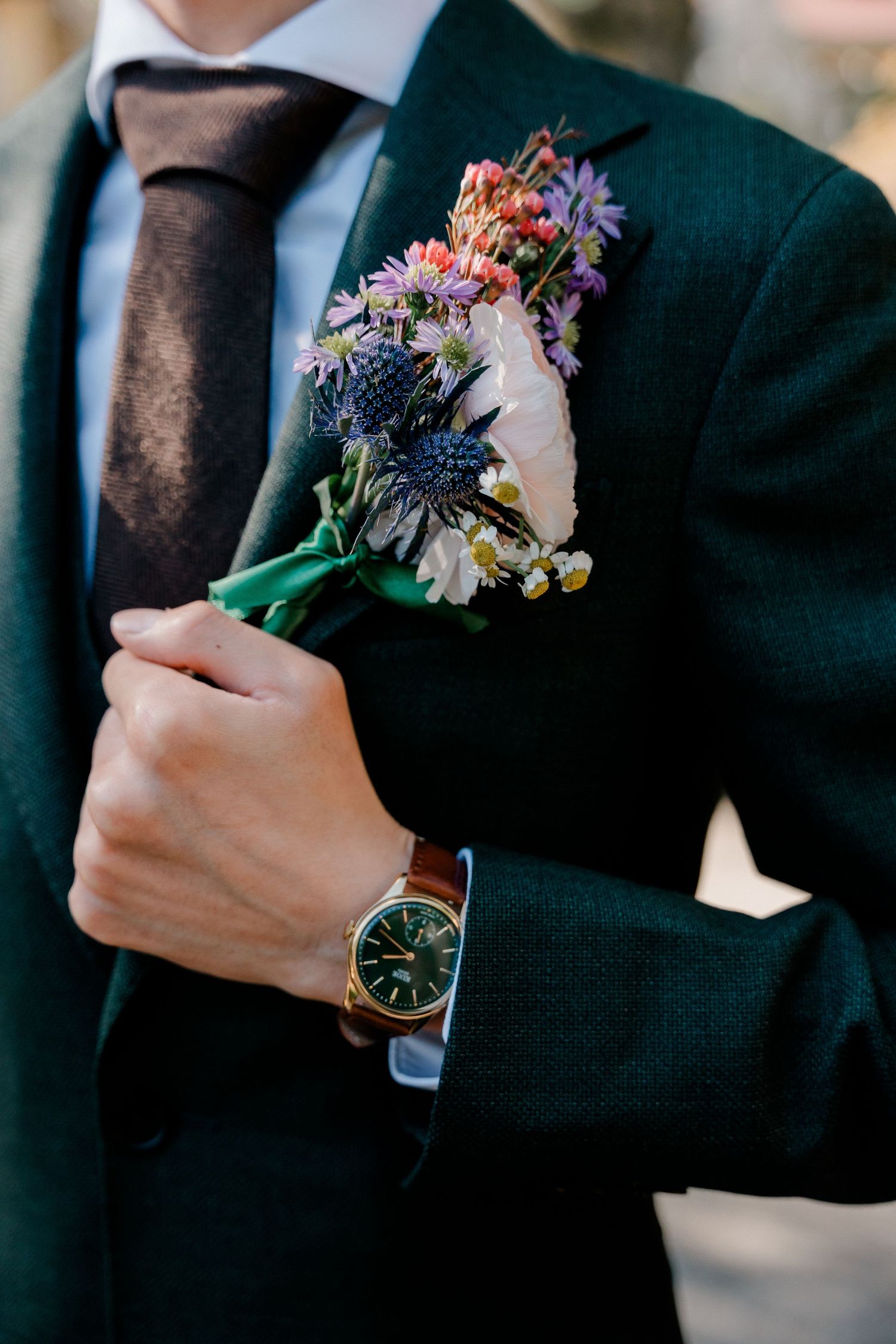 A close-up view of a black suit jacket with colorful floral boutonniere and wristwatch detail.