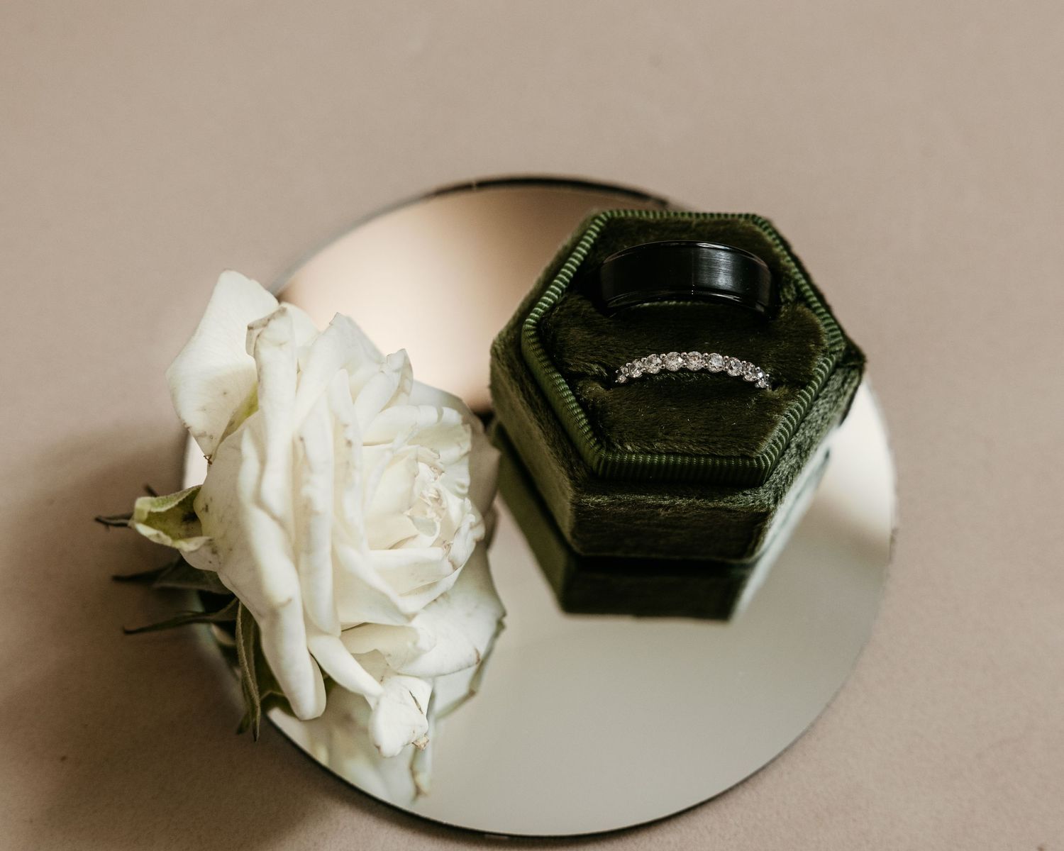 A white rose and dark hexagonal ring box reflected in a circular mirror on a neutral background.