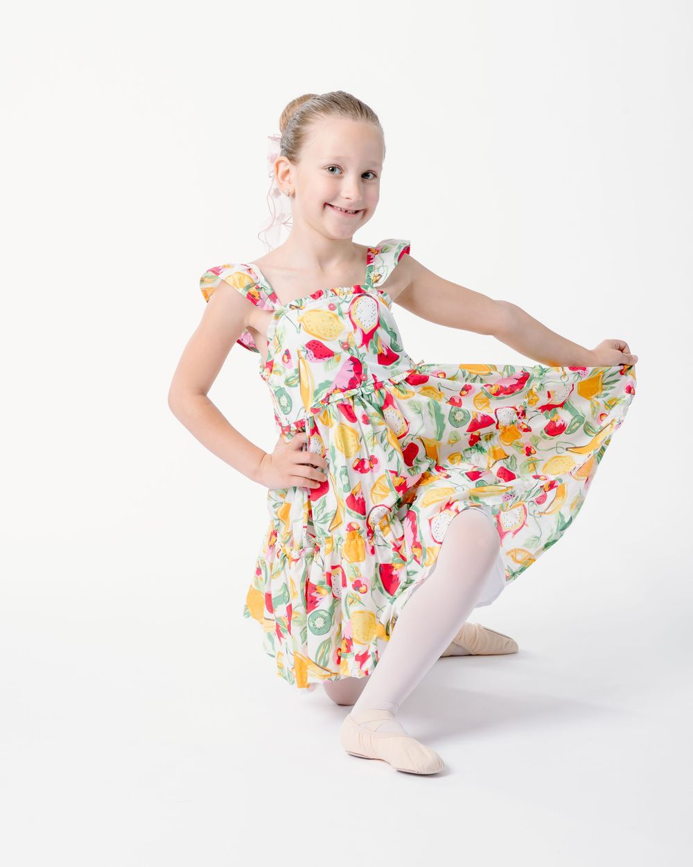Young dancer in floral print dress and white tights performs graceful ballet poses in studio against white background.