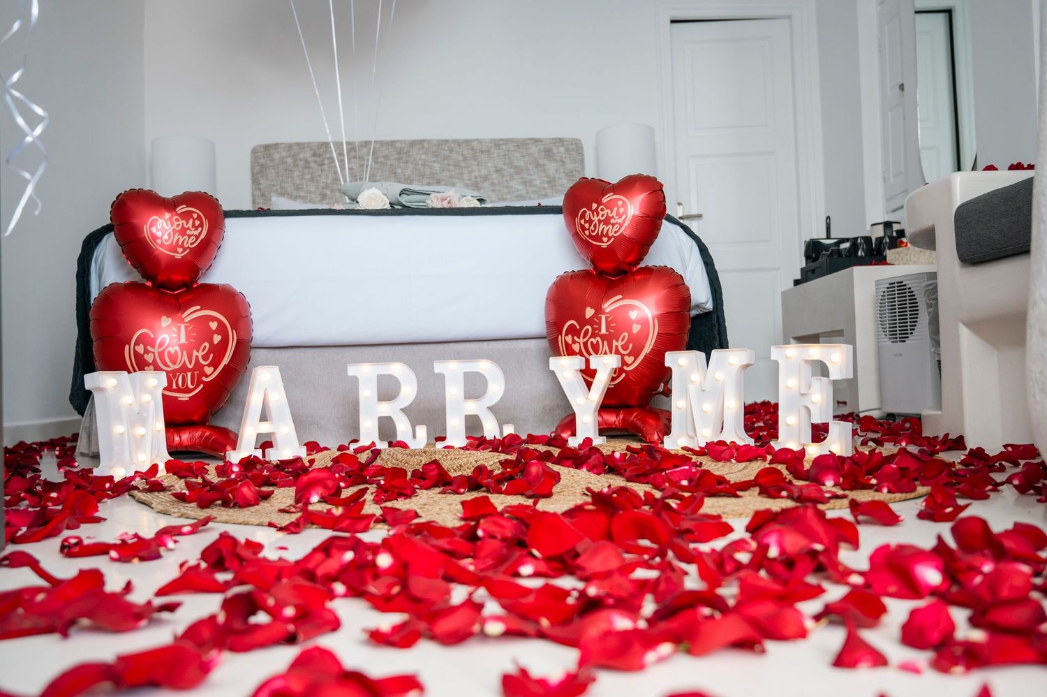 A marriage proposal setup with white 'MARRY ME' letters surrounded by red rose petals and heart-shaped balloons on a bed.