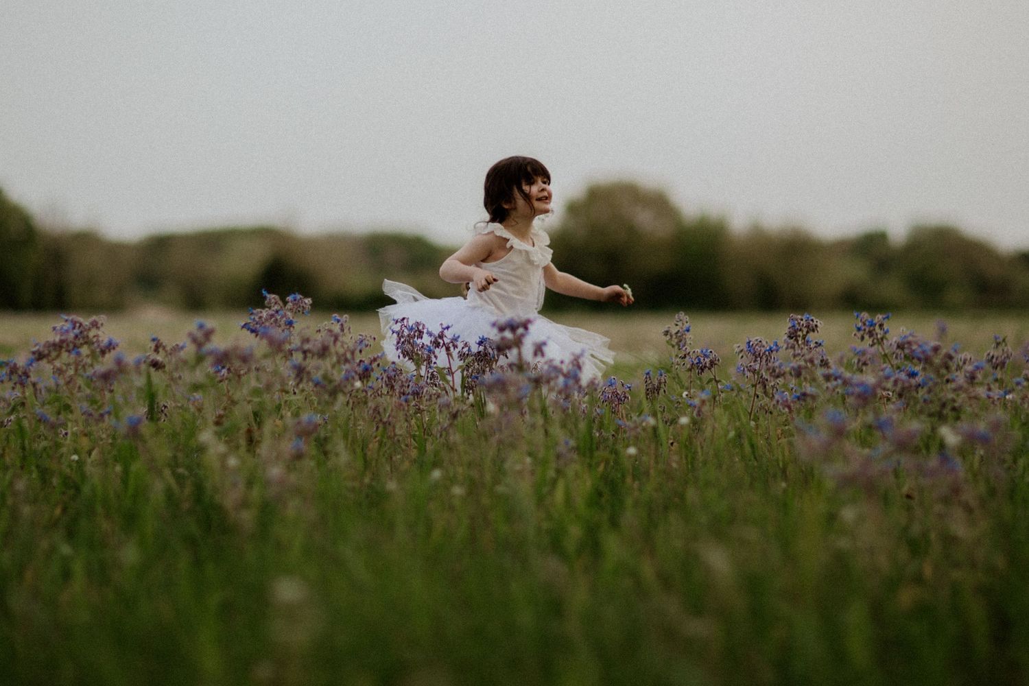 Child in white dress runs through purple wildflower field at dusk.