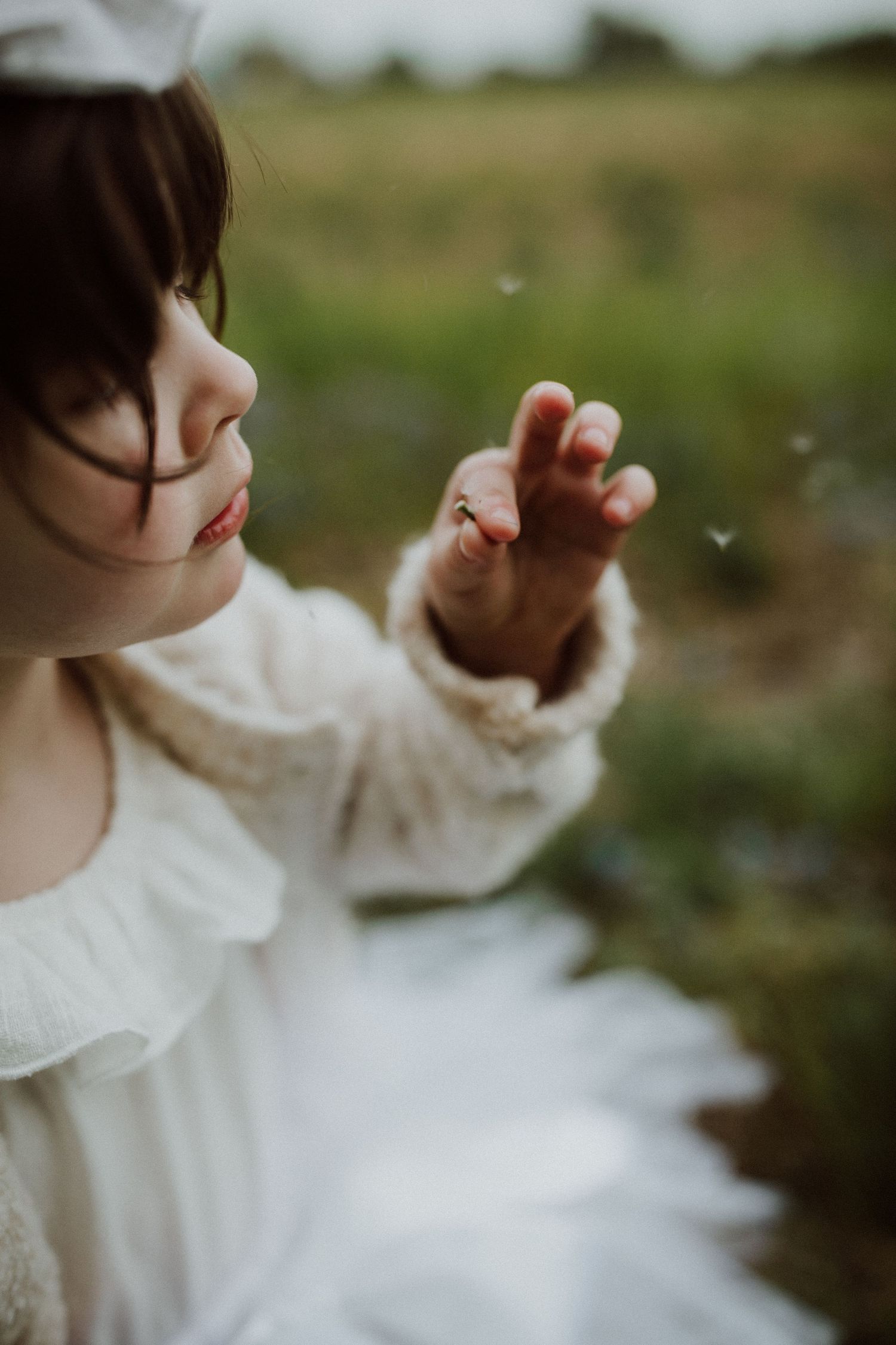 Child in white ruffled dress reaches for floating dandelion seeds.