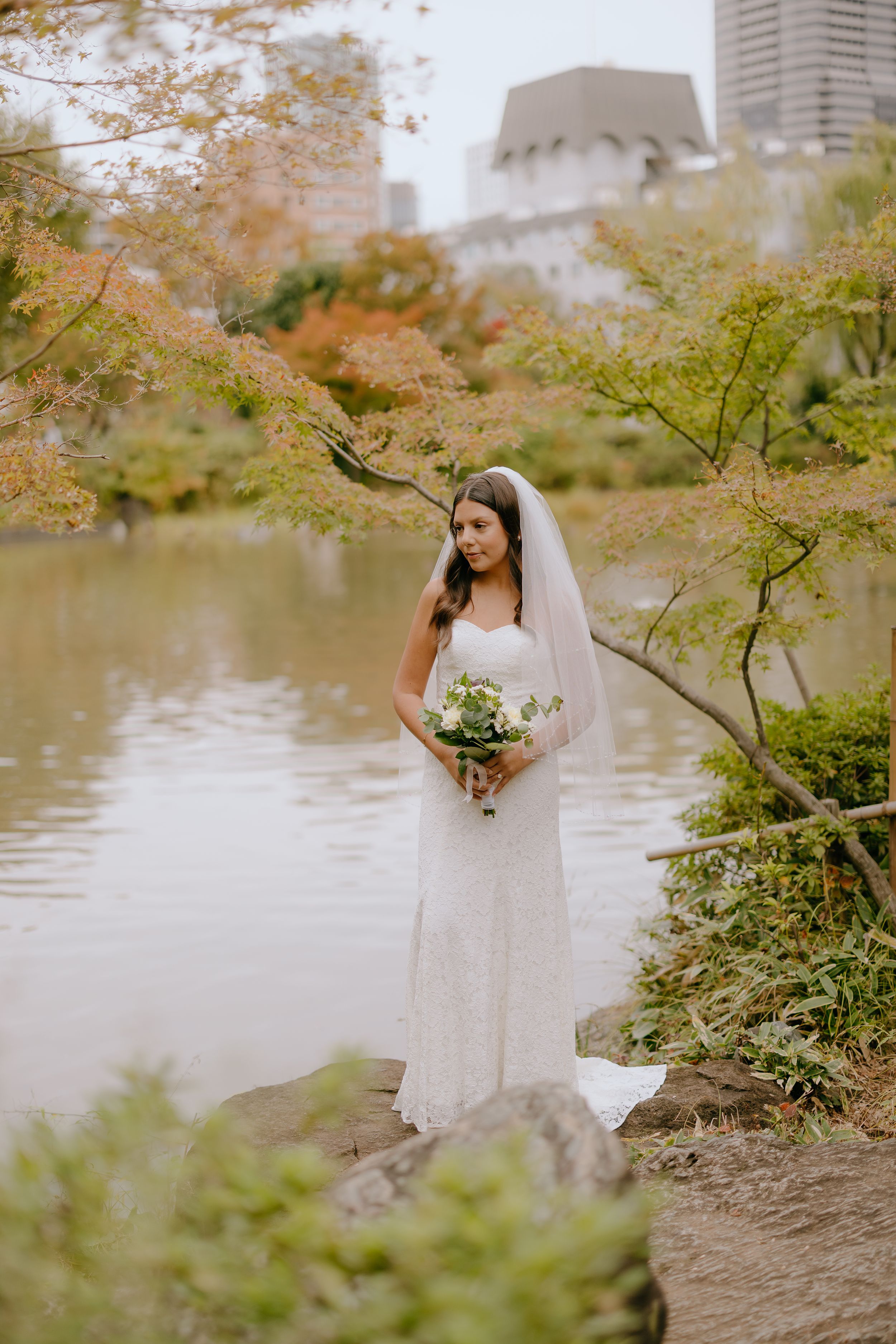 A bride in a white wedding dress poses gracefully by a lakeside surrounded by autumn foliage.
