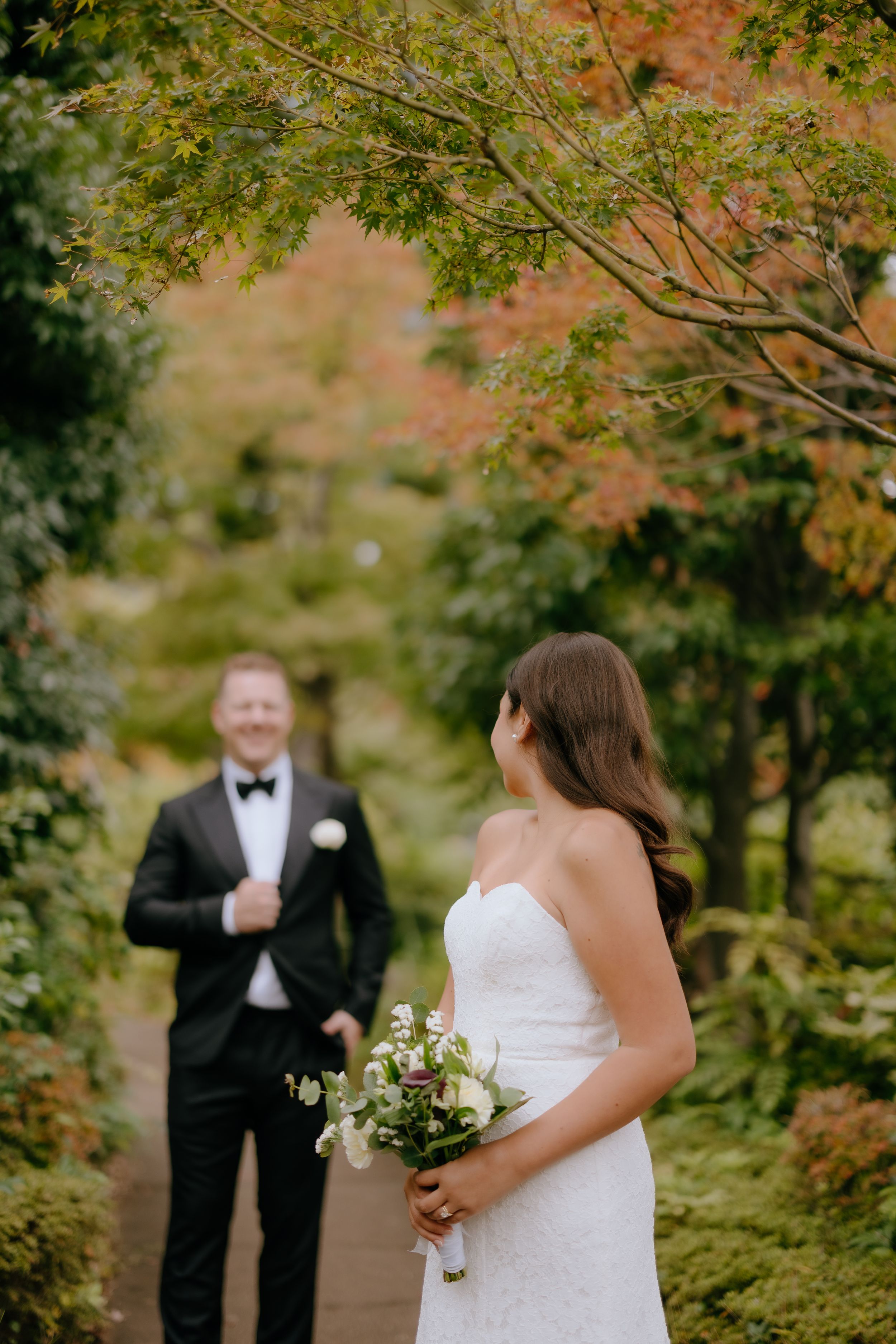 A wedding ceremony takes place under fall trees with the bride holding a white bouquet.