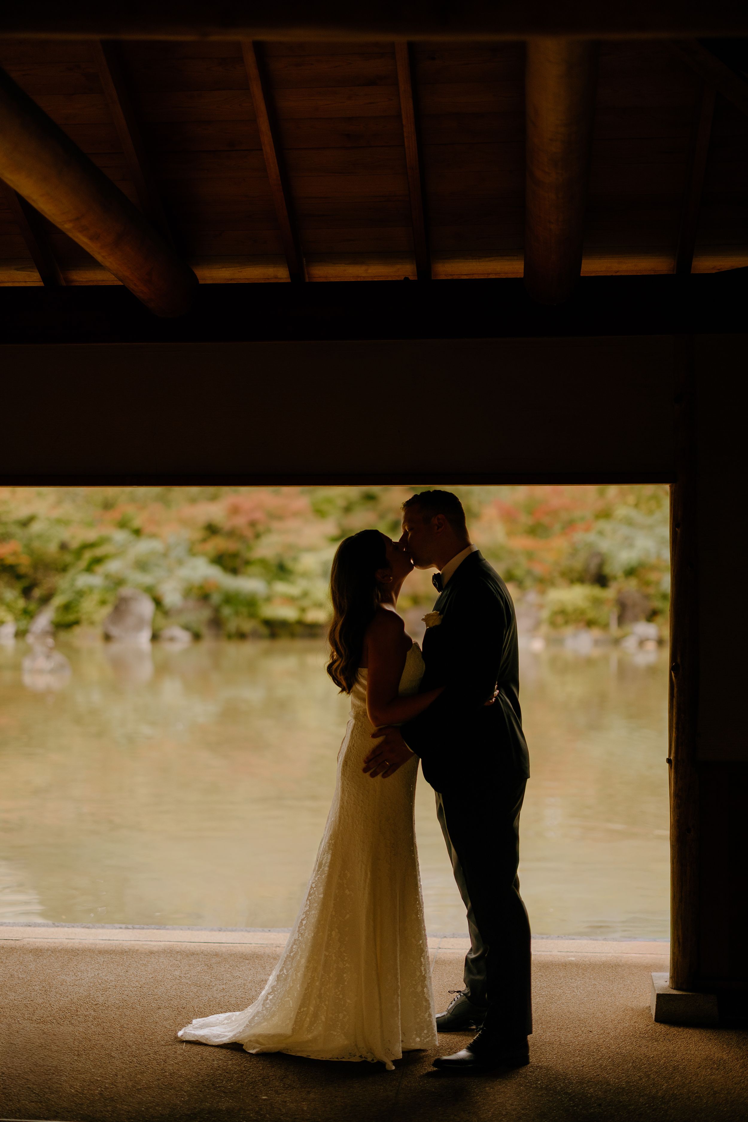 A romantic silhouette of a couple in wedding attire embracing under a covered area near a tranquil pond.