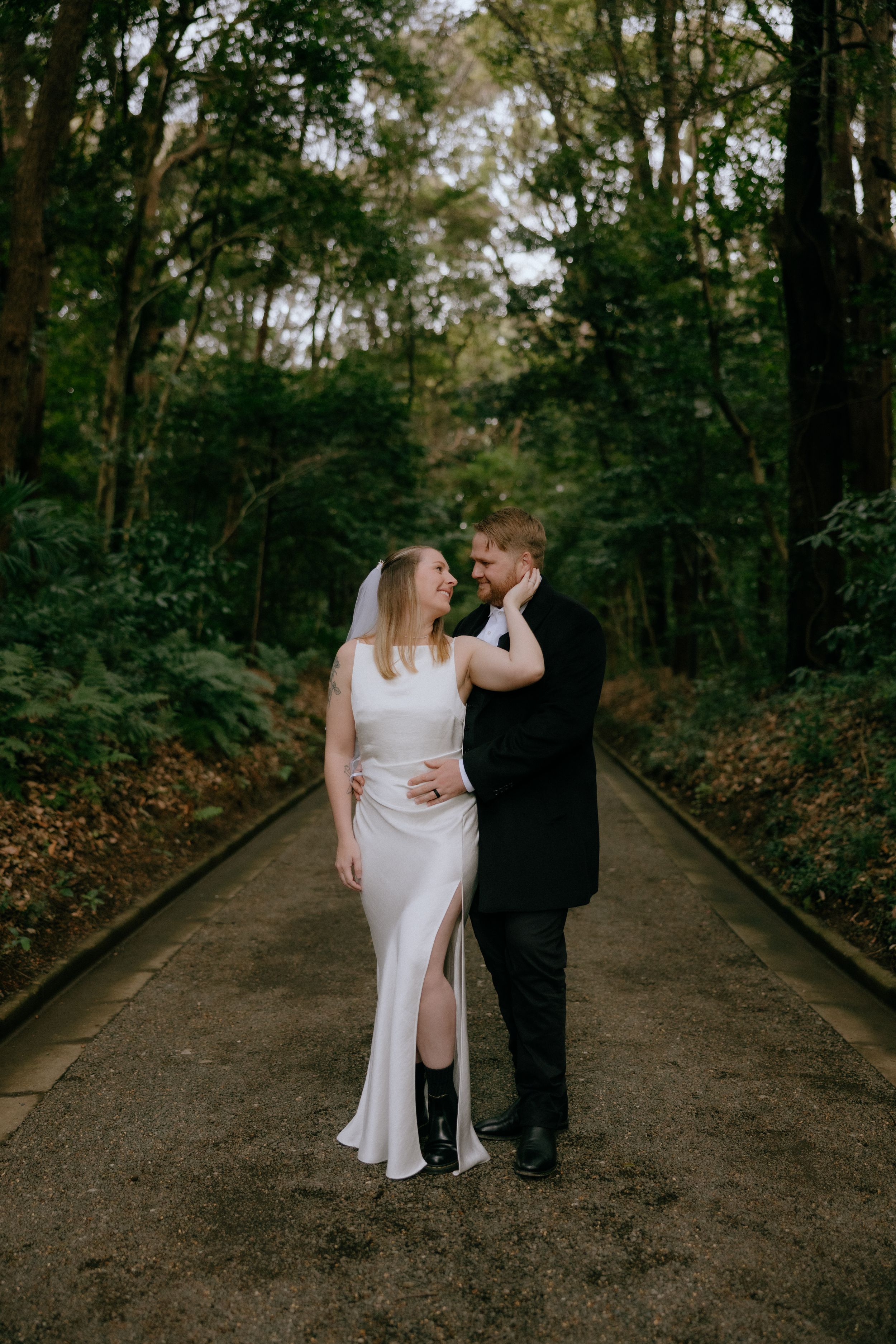 A couple in wedding attire share an intimate moment while walking along a tree-lined path in a forest setting.