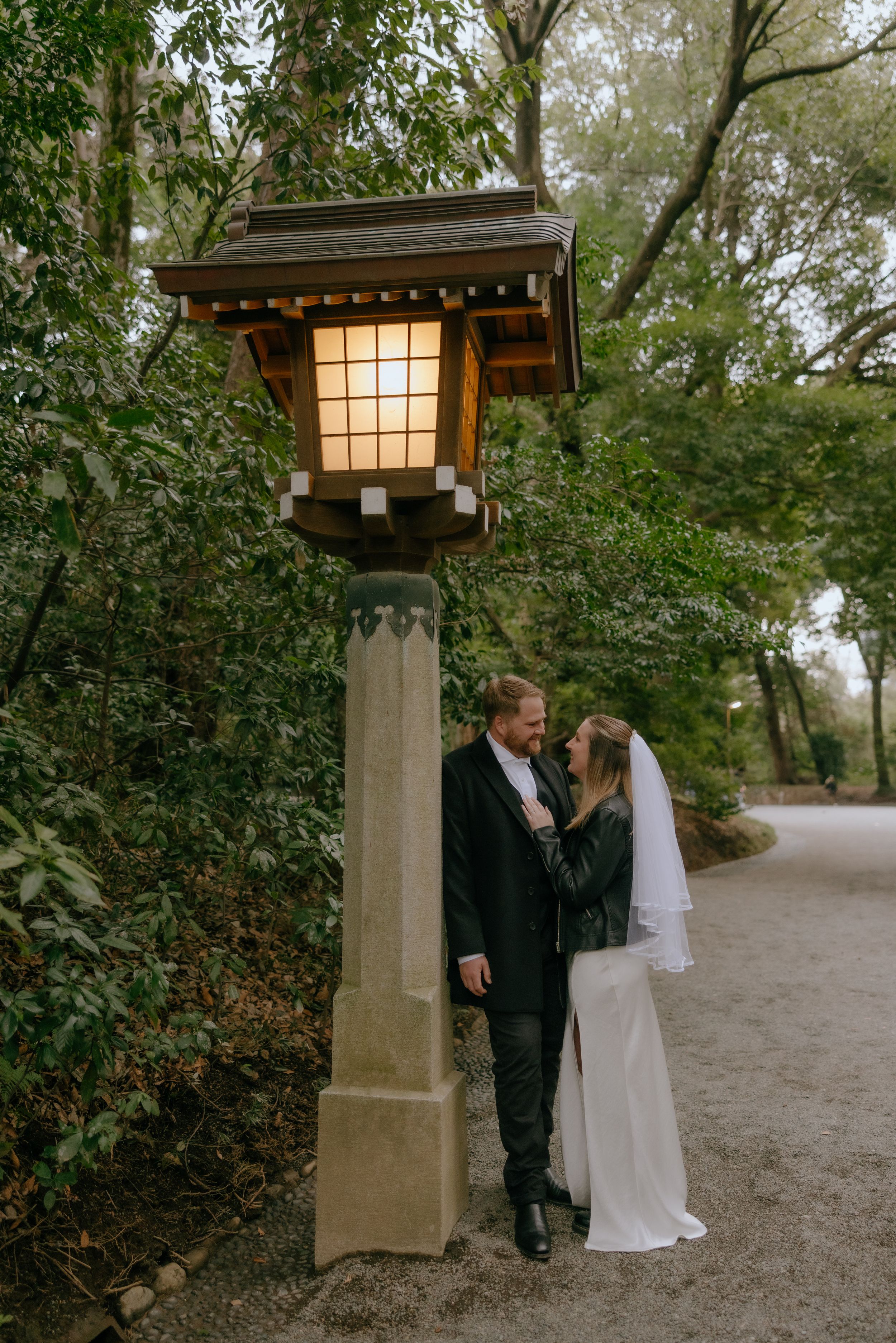 A couple in wedding attire stands beneath an illuminated Japanese lantern on a garden path during dusk.