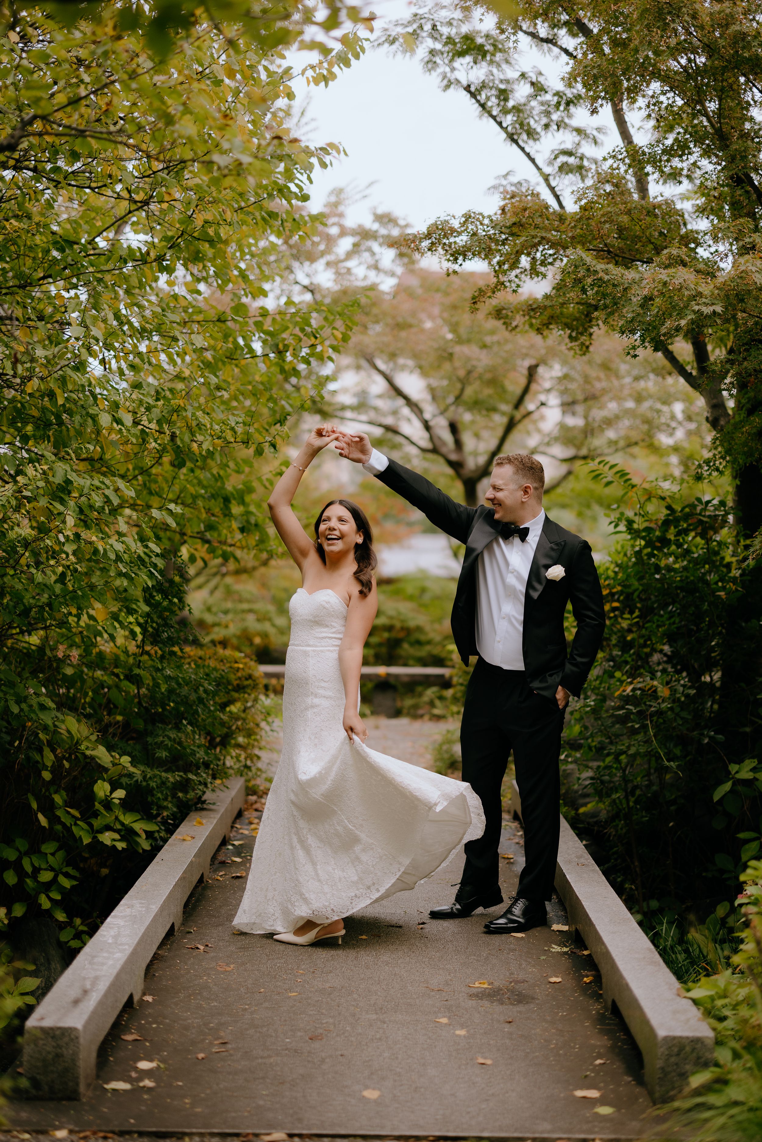 Joyful couple in wedding attire dance and twirl playfully on a garden bridge surrounded by autumn foliage.