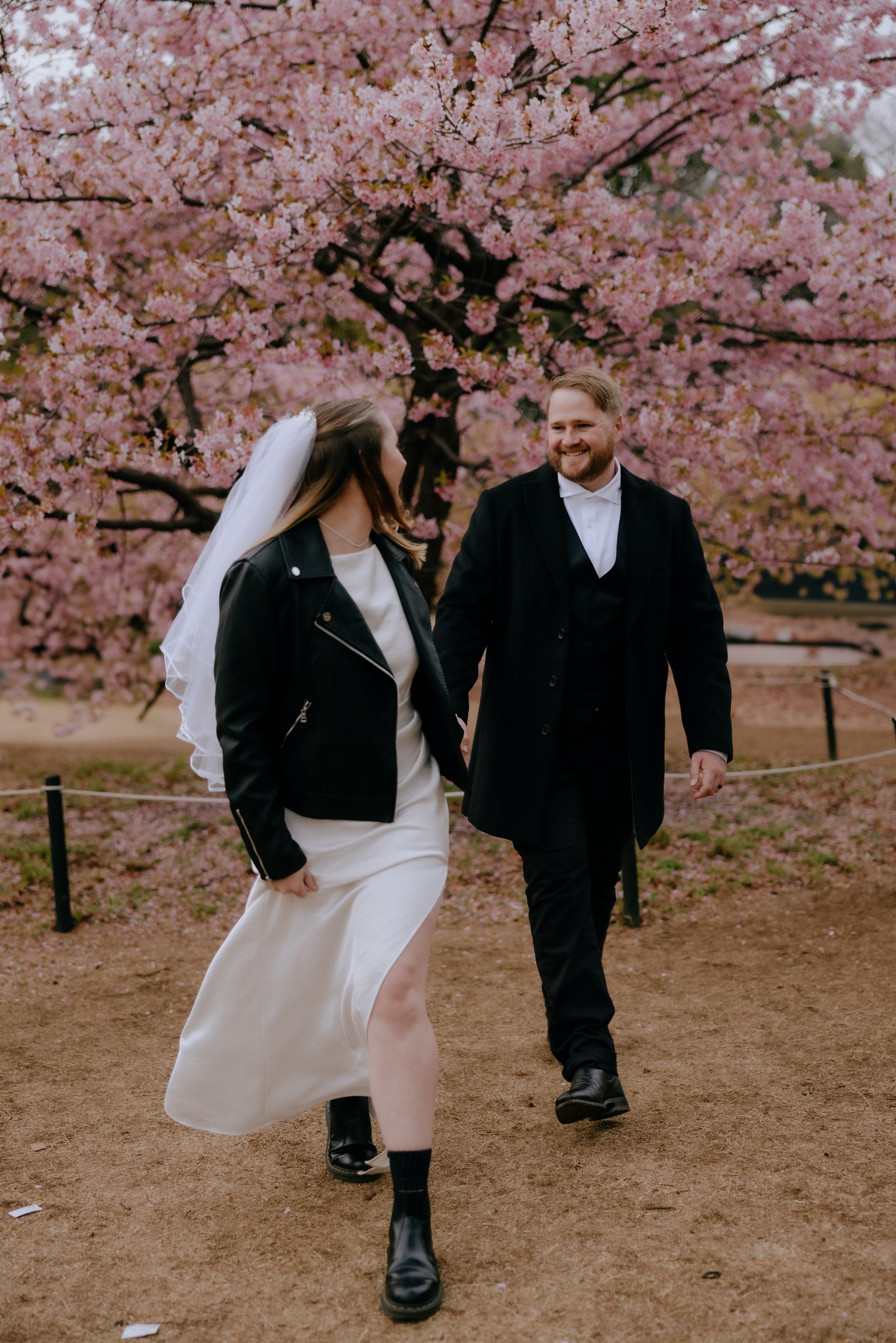 A couple in wedding attire pose playfully under blooming pink cherry blossom trees on a spring day.